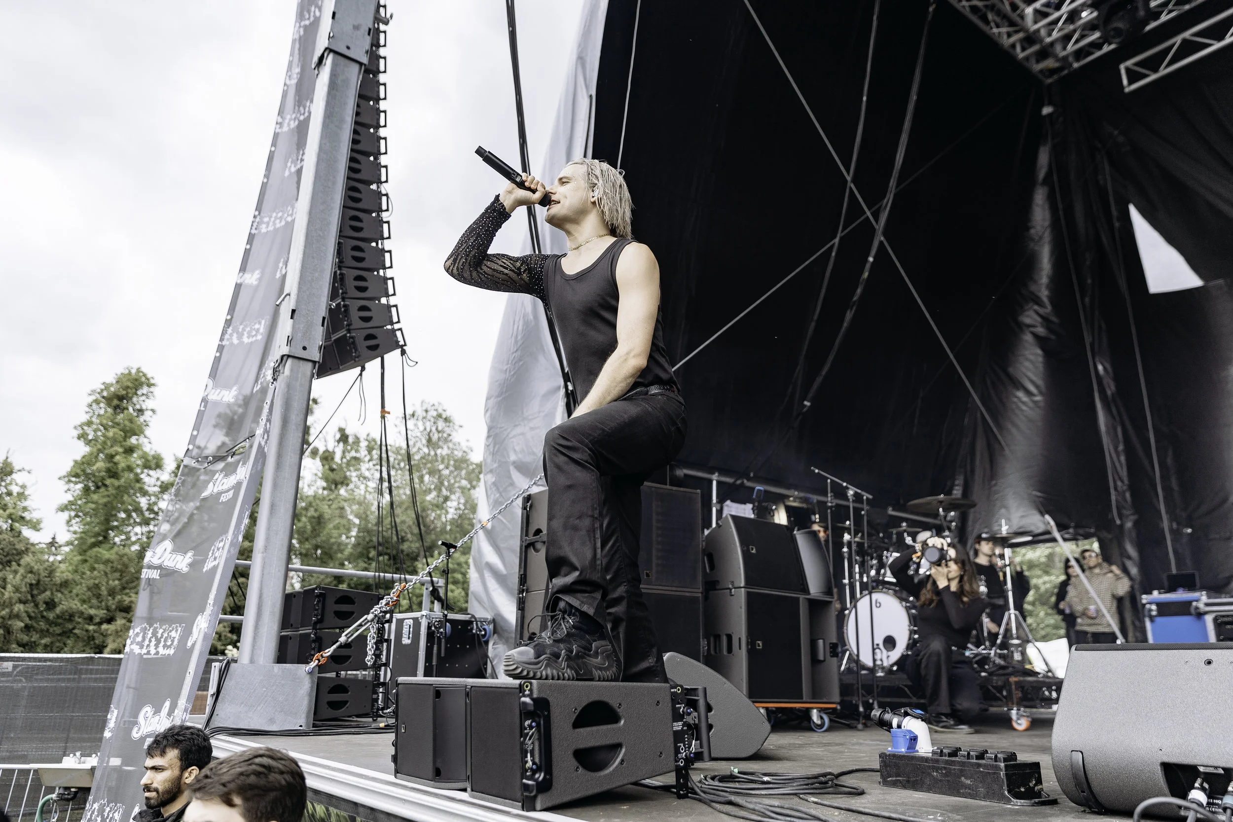 A musician with platinum blonde hair singing into a microphone on an outdoor stage, wearing a sleeveless black top and black pants. There are stage equipment, a drummer, and photographers in the background. Trees are visible in the distance under a c