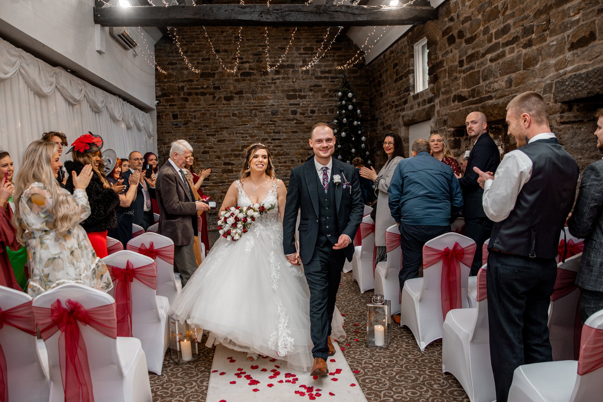 Bride and groom walking down the aisle after wedding ceremony, surrounded by seated guests, in a decorated indoor venue with stone walls, Christmas tree, and string lights.