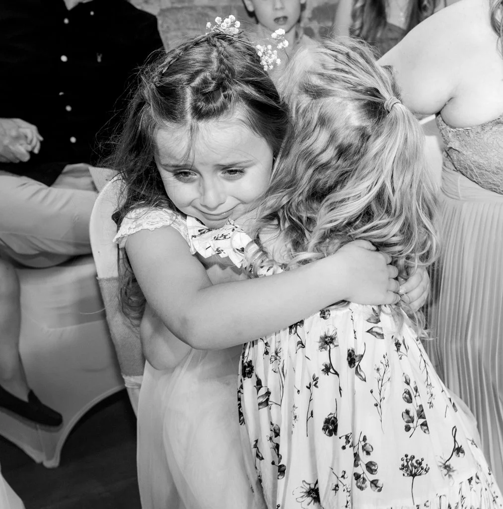 Two young girls hugging at a gathering, one visibly emotional, in black and white.