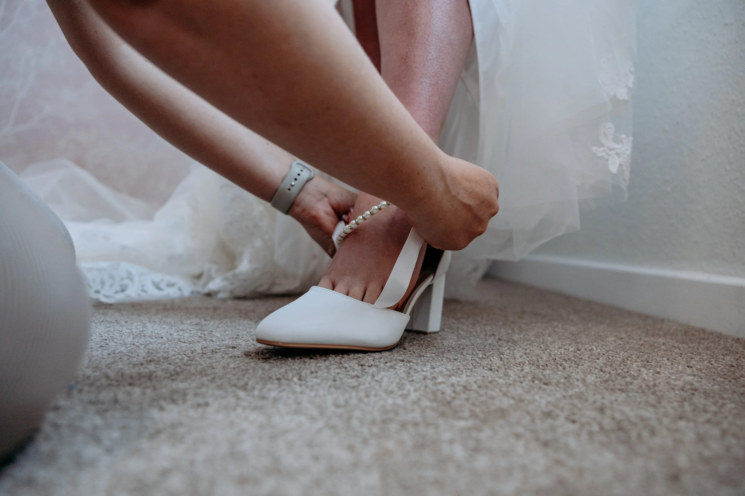 A woman in a wedding dress adjusting a white high-heeled shoe with ankle straps and a pearl bracelet on her ankle.