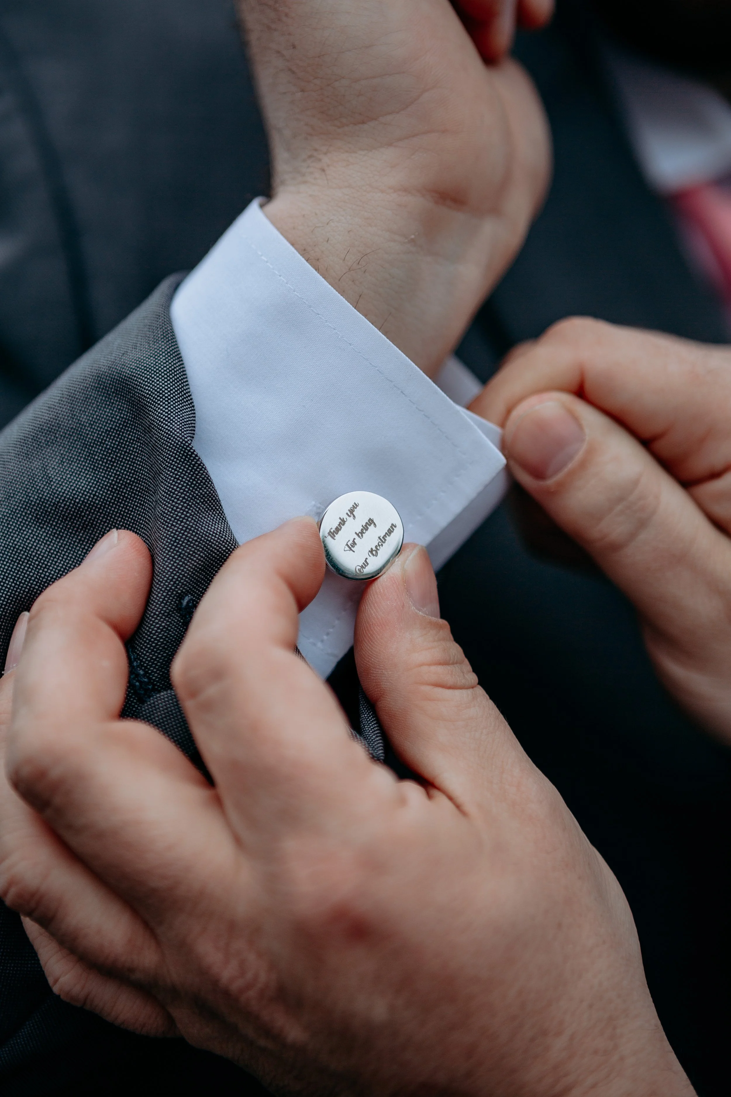 Someone is pinning a wedding boutonniere with a small round badge that reads 'Thank you for being our witness' onto a man's suit jacket near his tuxedo shirt collar.