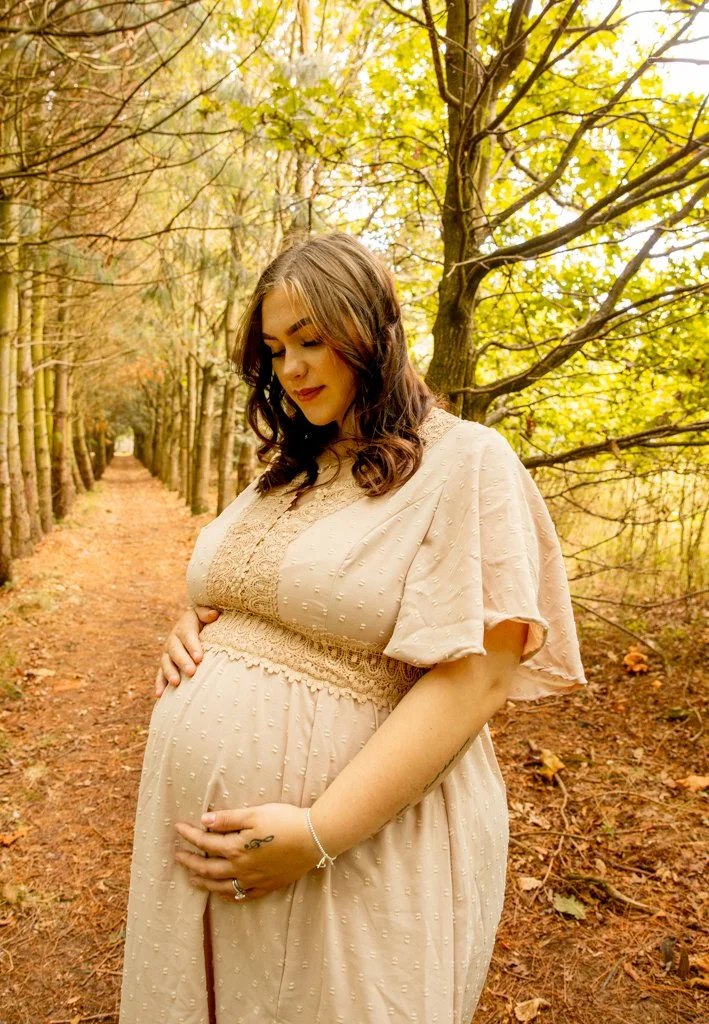 A pregnant woman in a beige dress standing in a wooded area with autumn leaves on the ground and trees with yellow leaves.
