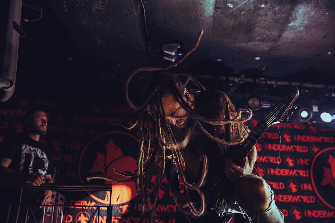 A musician with dreadlocks playing an electric guitar on stage during a concert with a band member in the background, in front of a backdrop with the word 'Underworld' repeated multiple times.