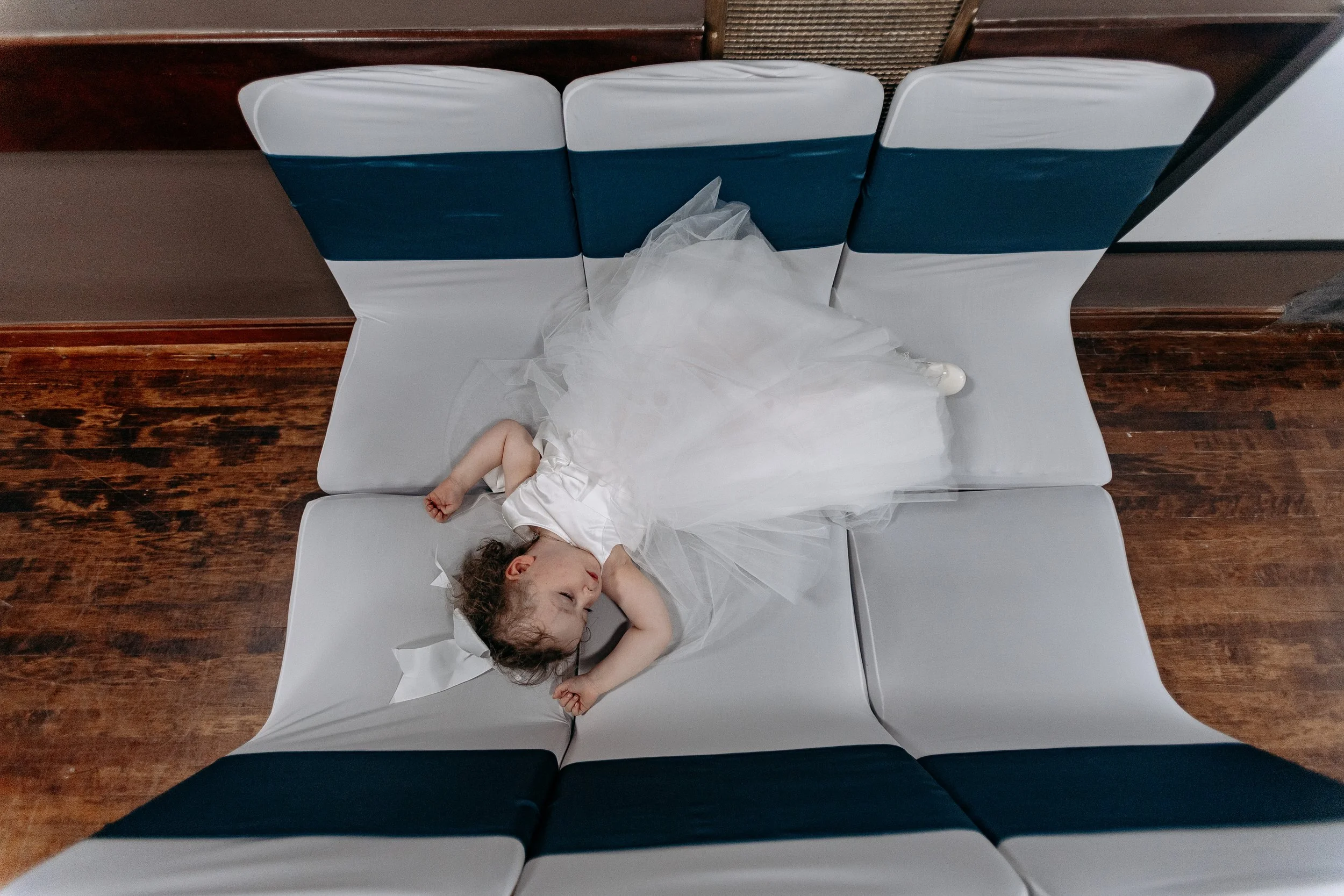 A young girl in a white dress with a tulle skirt is lying on a row of white and blue cushioned chairs, sleeping with her hands over her head and her face turned to the side.