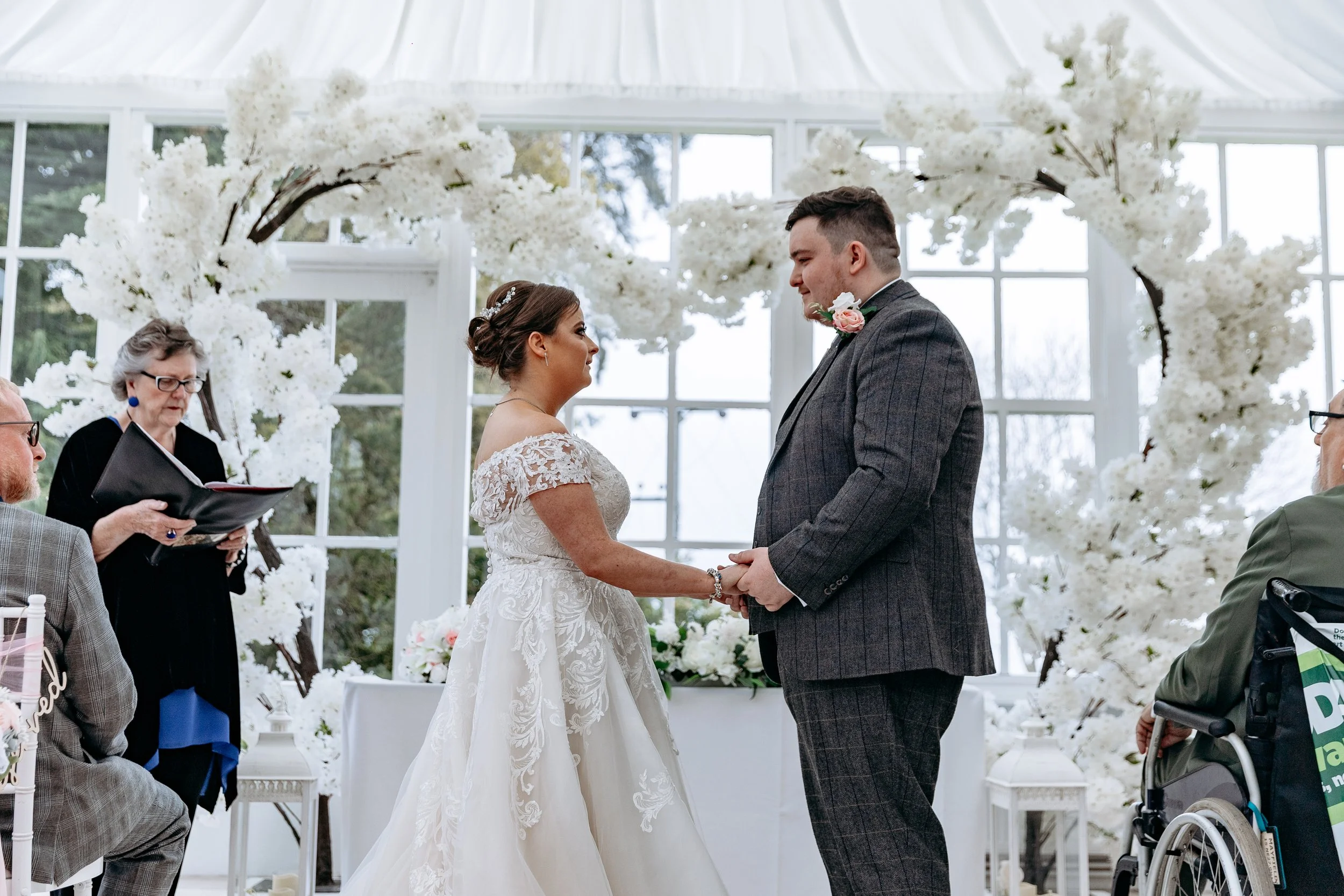 Bride and groom holding hands during their wedding ceremony in a glass-enclosed venue decorated with white flowers.