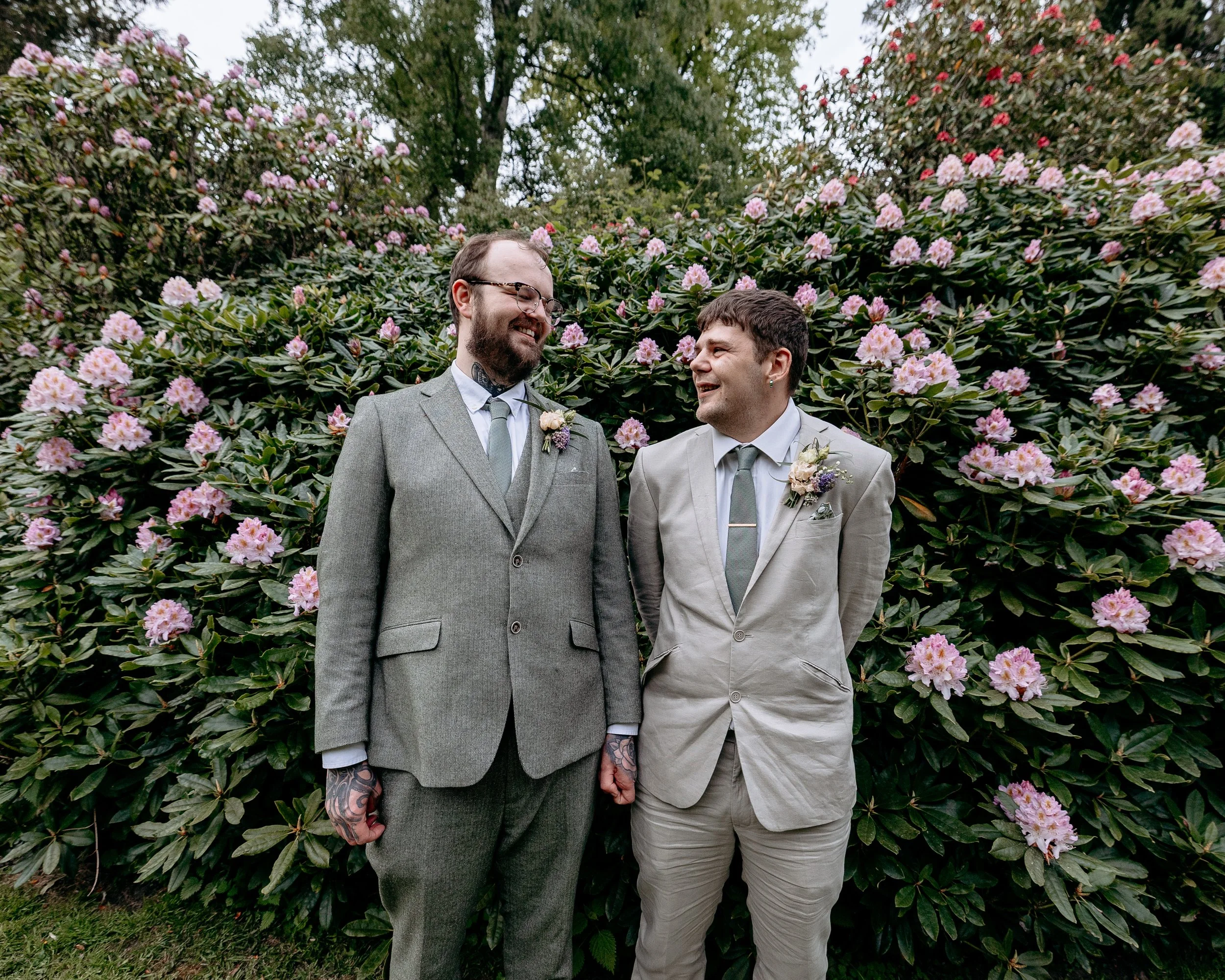 Two men dressed in suits standing in front of a bush with pink flowers, holding hands, smiling and looking at each other during a wedding or special event.