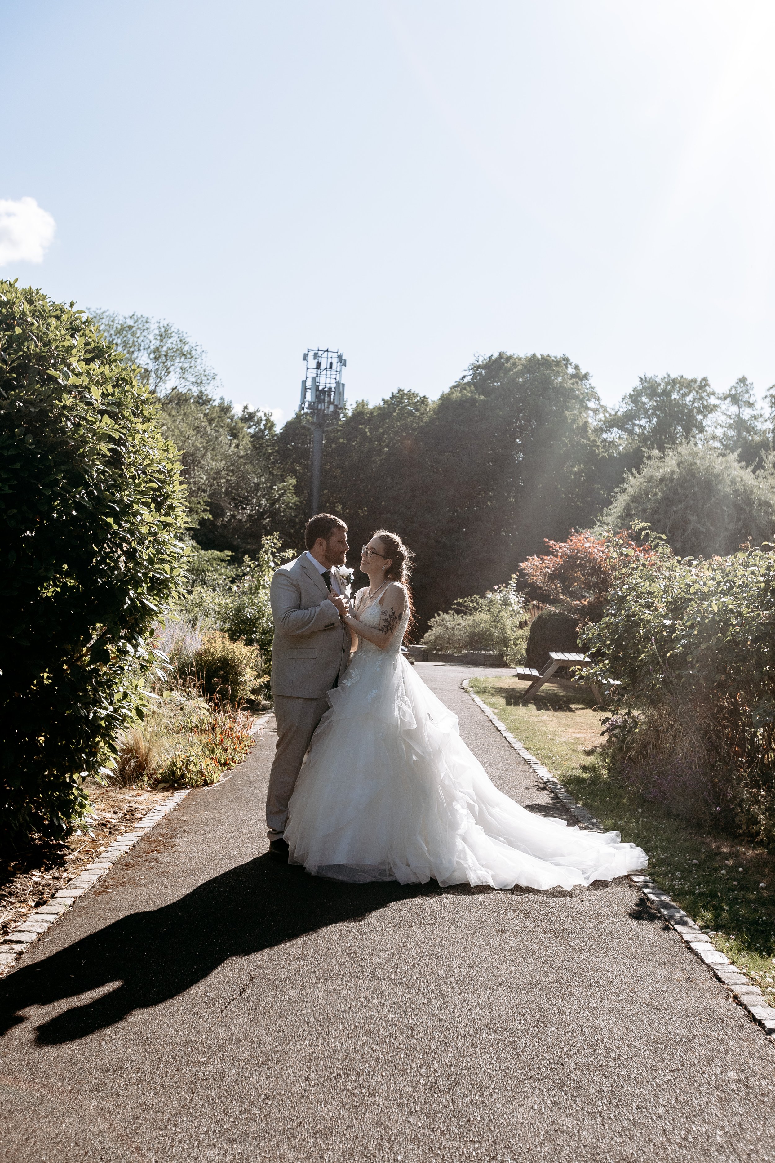 A bride and groom standing together on a park pathway, surrounded by greenery and sunlight, sharing a moment.