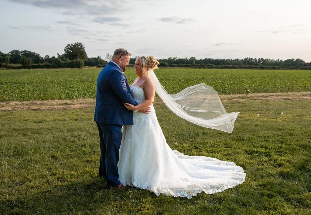 A bride and groom in wedding attire standing close together in a grassy field, smiling at each other, with trees and a cloudy sky in the background.