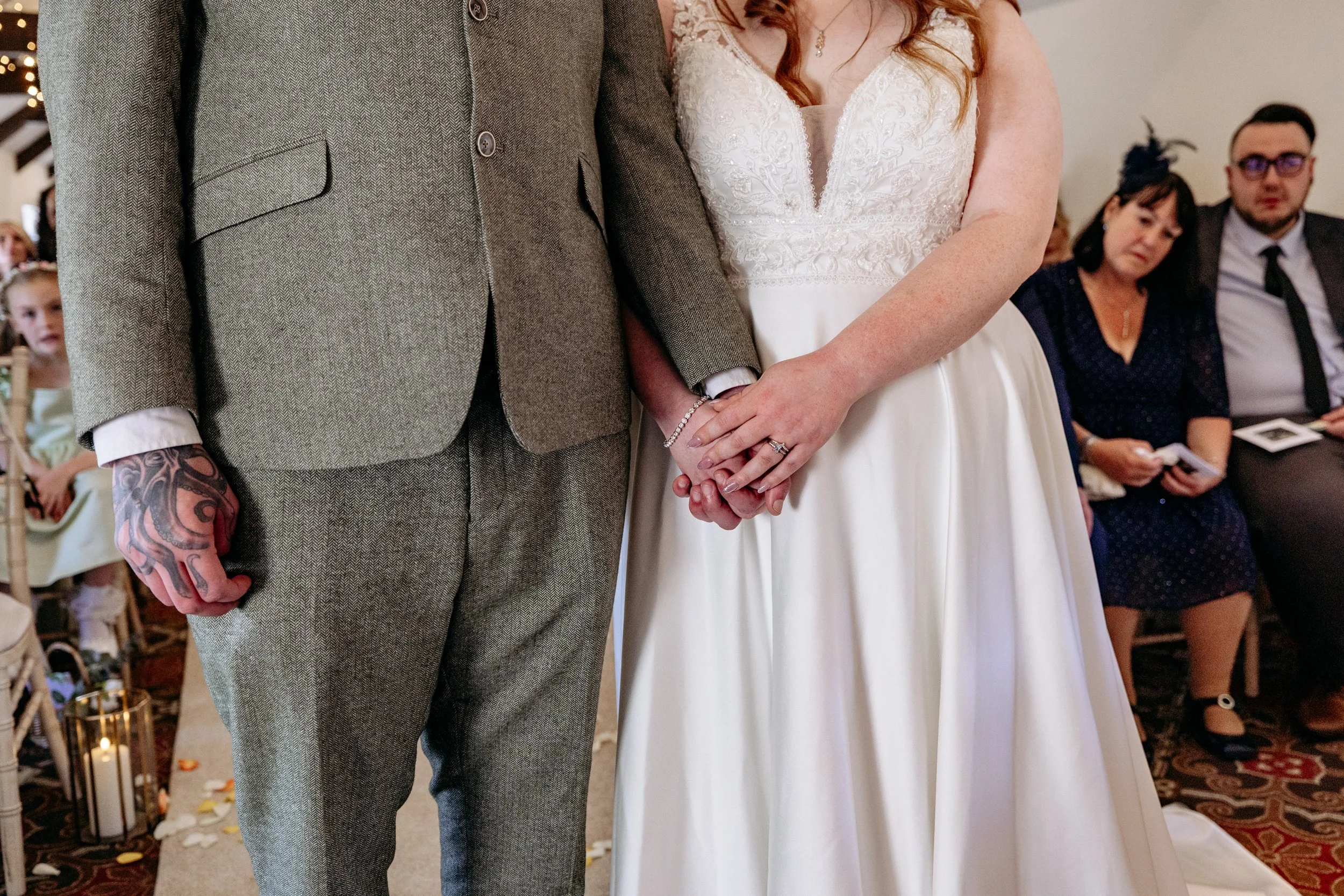 Bride and groom holding hands during a wedding ceremony, with wedding guests in the background.