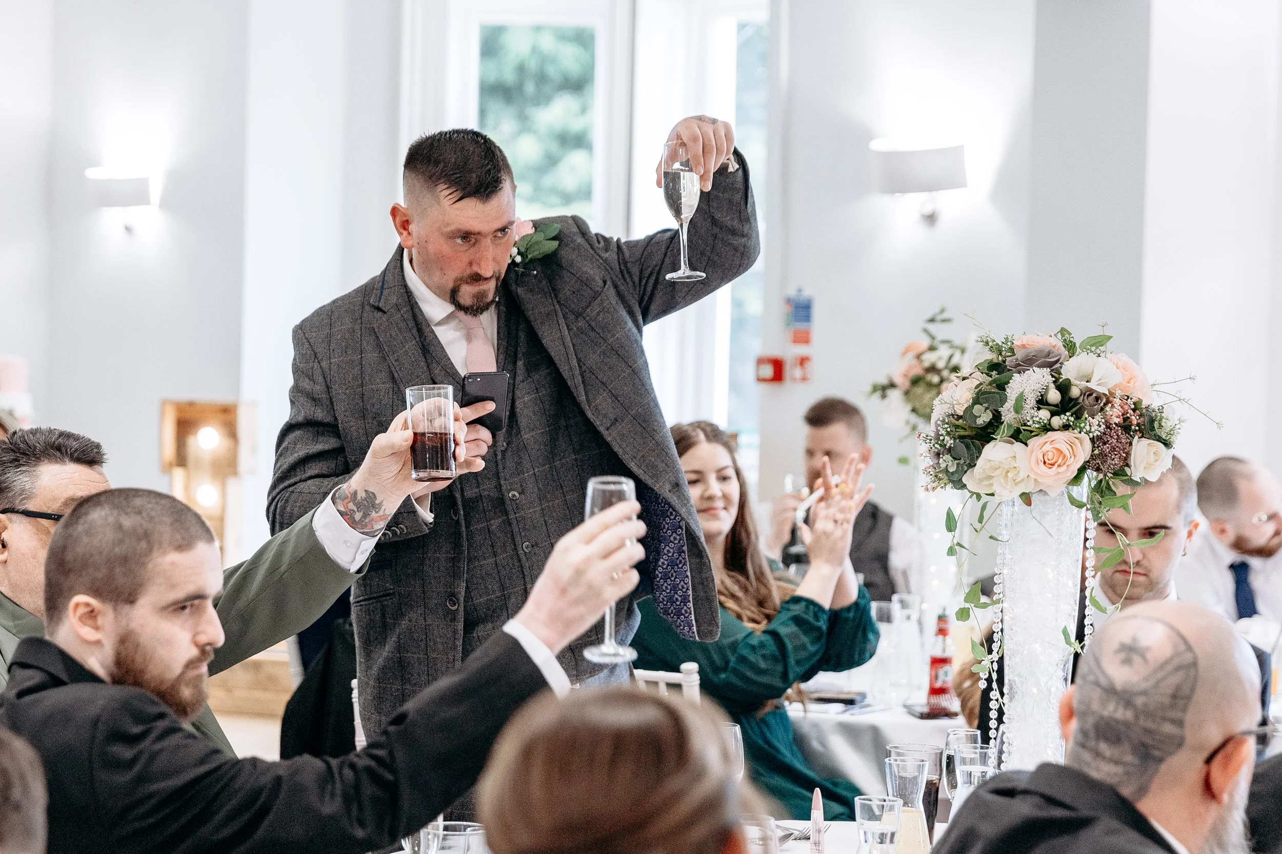 Man making a toast at a wedding reception, holding a glass of champagne, while others seated around him raise their glasses.