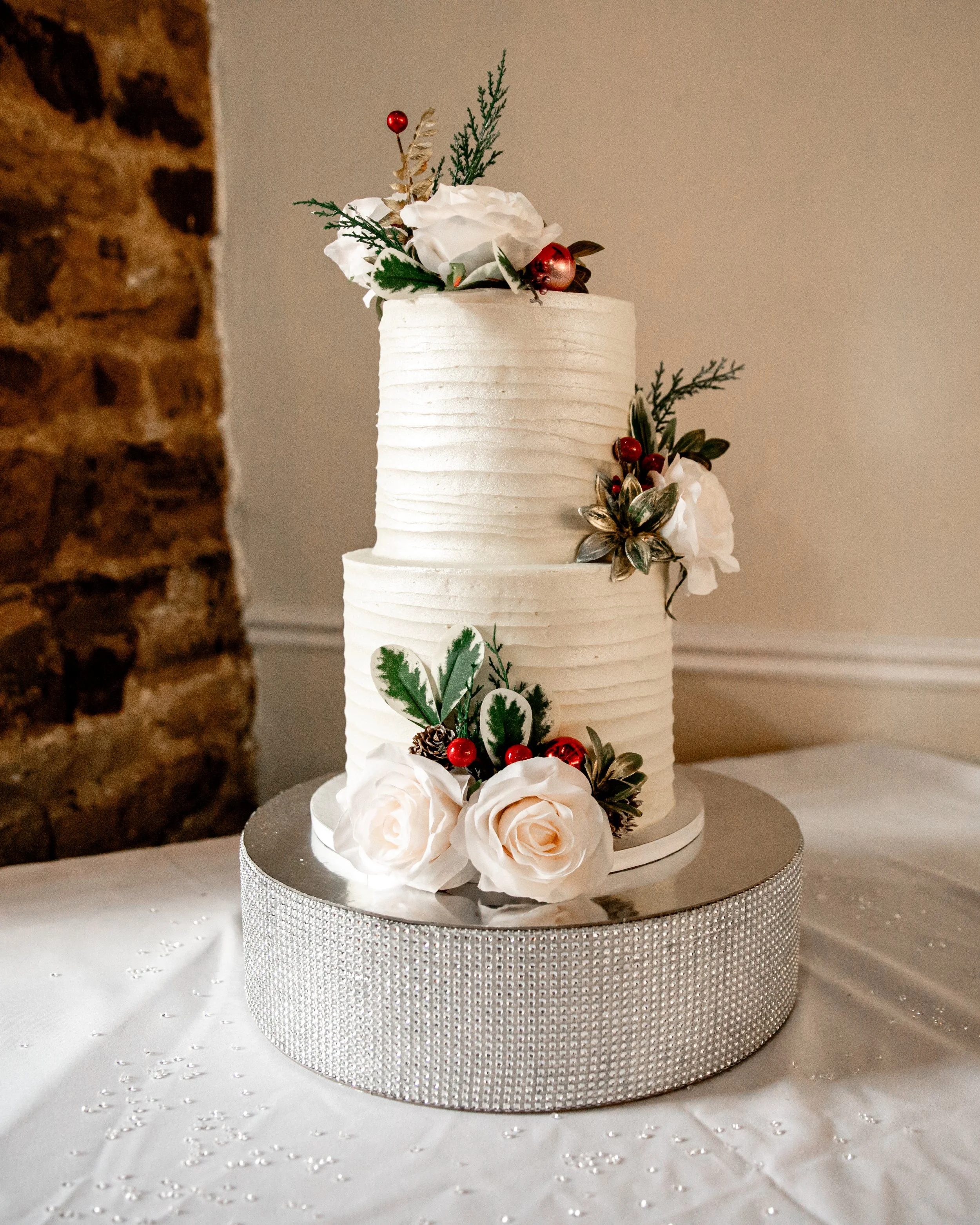 A tall white wedding cake decorated with white roses, greenery, red berries, and gold-foiled leaves on a silver, textured cake stand.