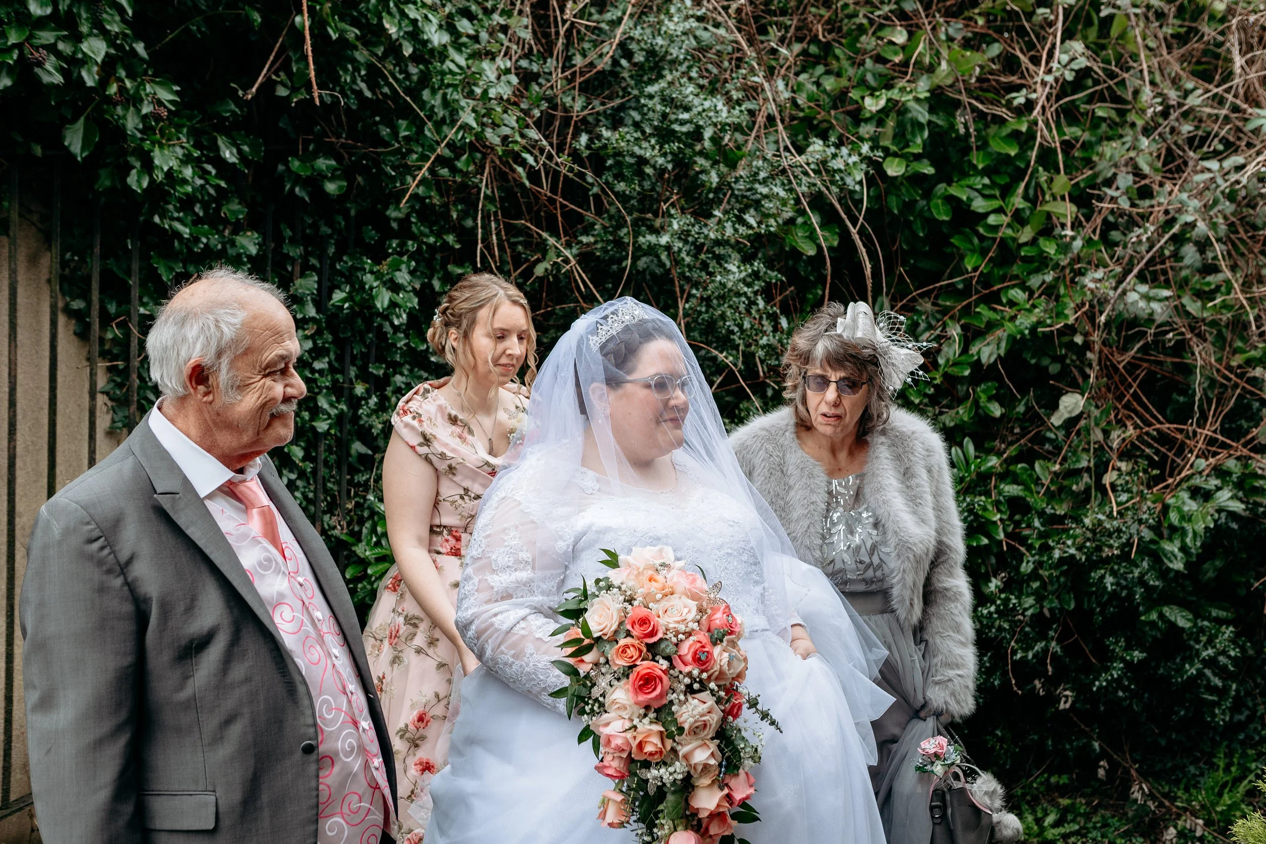 A bride holding a bouquet of pink and cream roses standing with four older adults, in a garden with green foliage.
