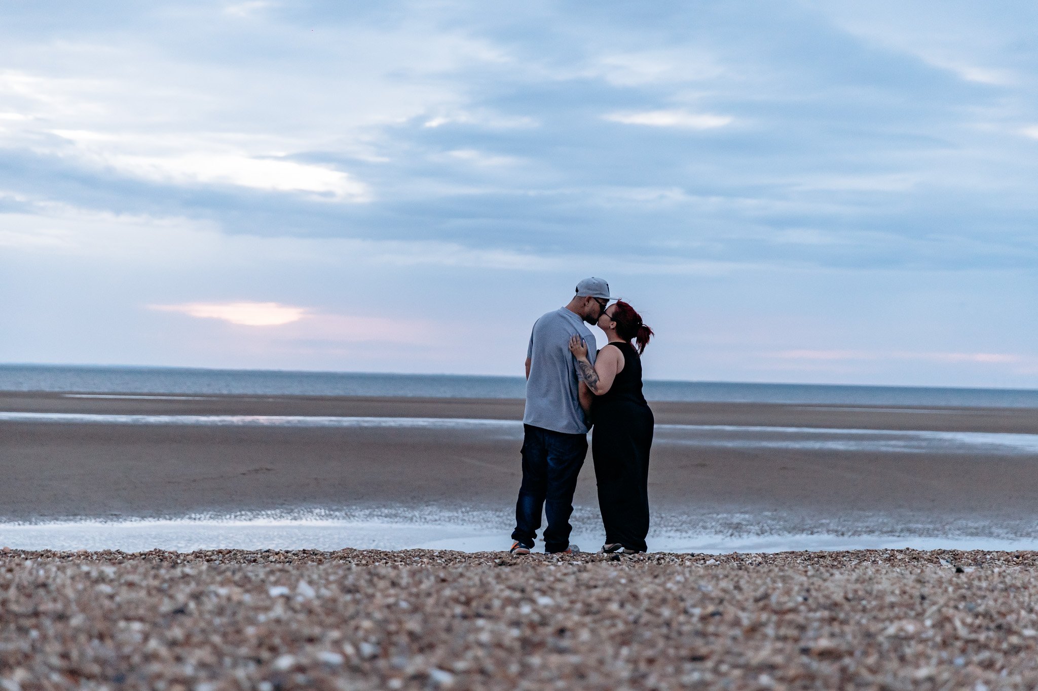 A couple kissing on the beach during sunset, with the ocean and cloudy sky in the background.