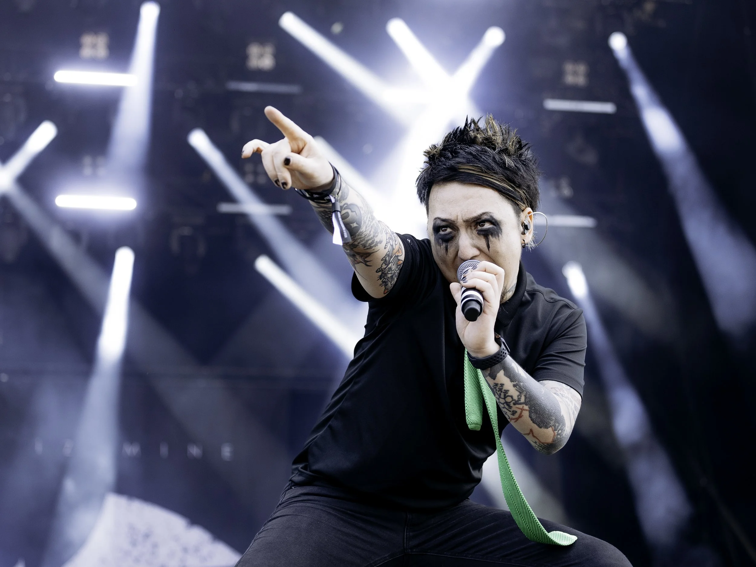 A young female singer with tattoos and black makeup on her face singing into a microphone on stage with bright white stage lights shining behind her.