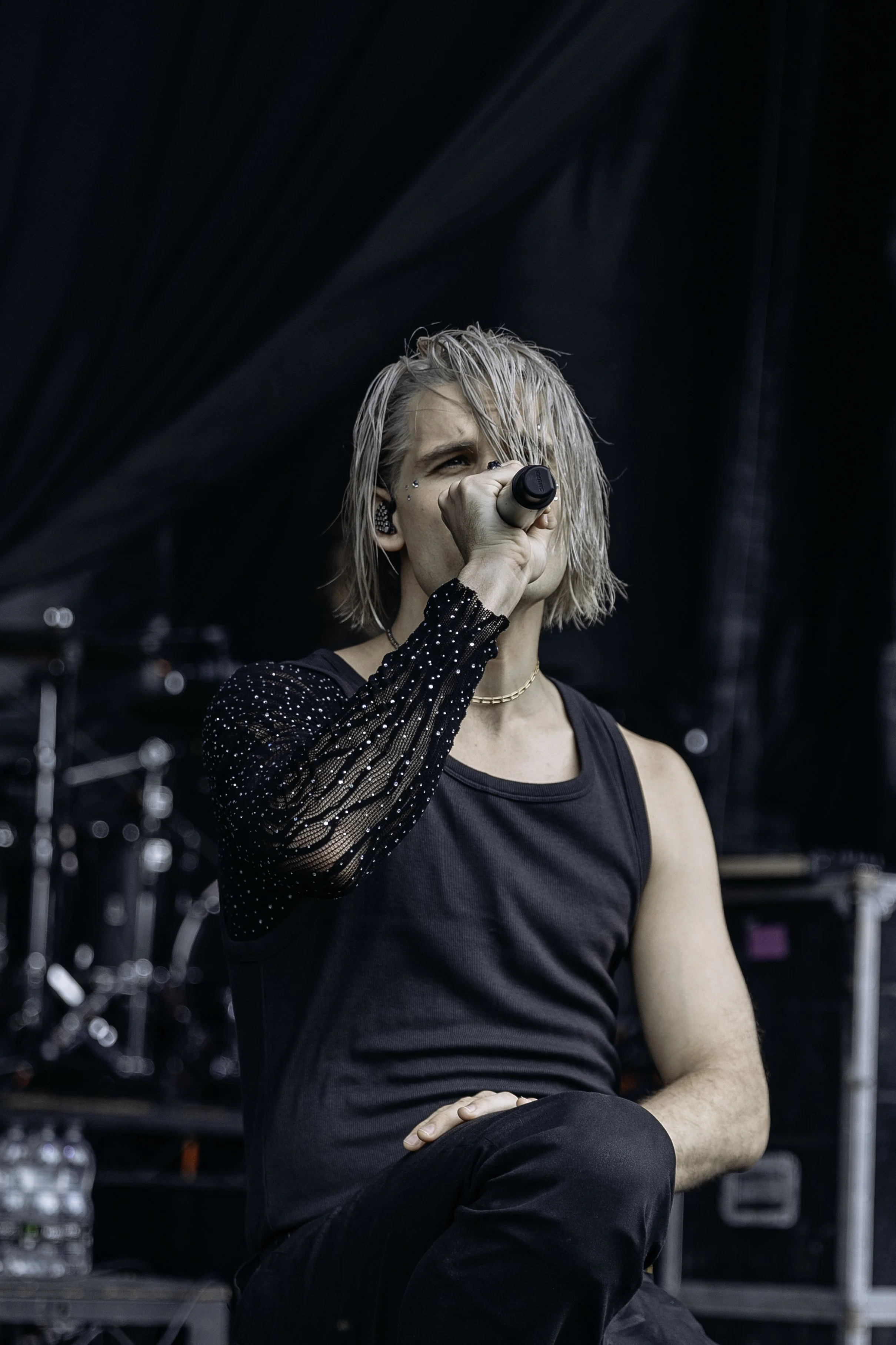 A male singer with platinum blonde hair holding a microphone while performing on stage, wearing a black sleeveless top with mesh sleeves decorated with small beads.
