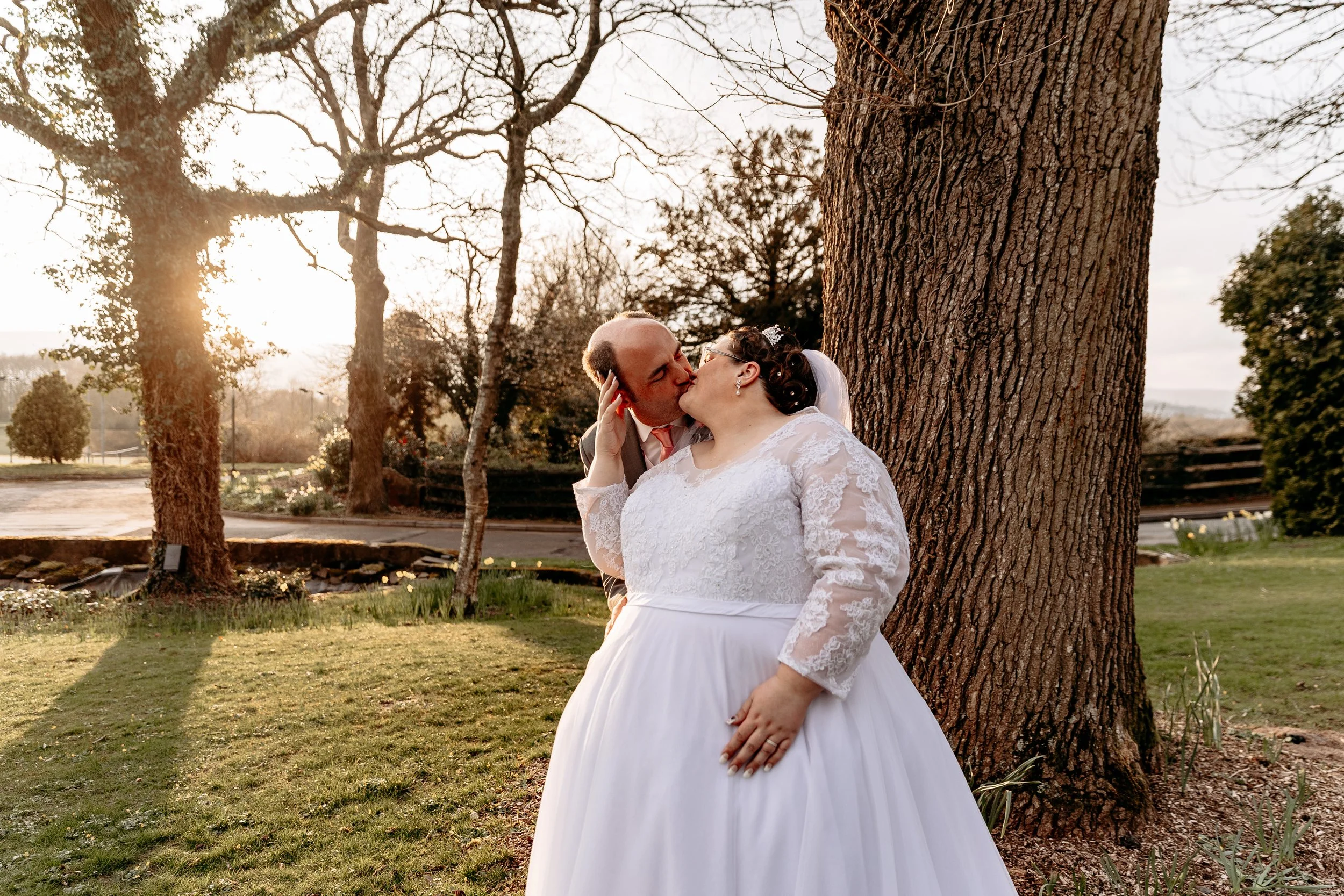A bride and groom kissing outdoors by a large tree during sunset.