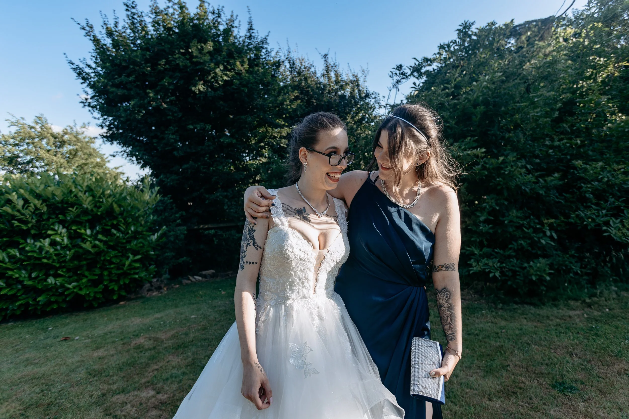 Two women, one in a white wedding dress and the other in a dark blue dress, share a joyful moment outdoors with greenery in the background.