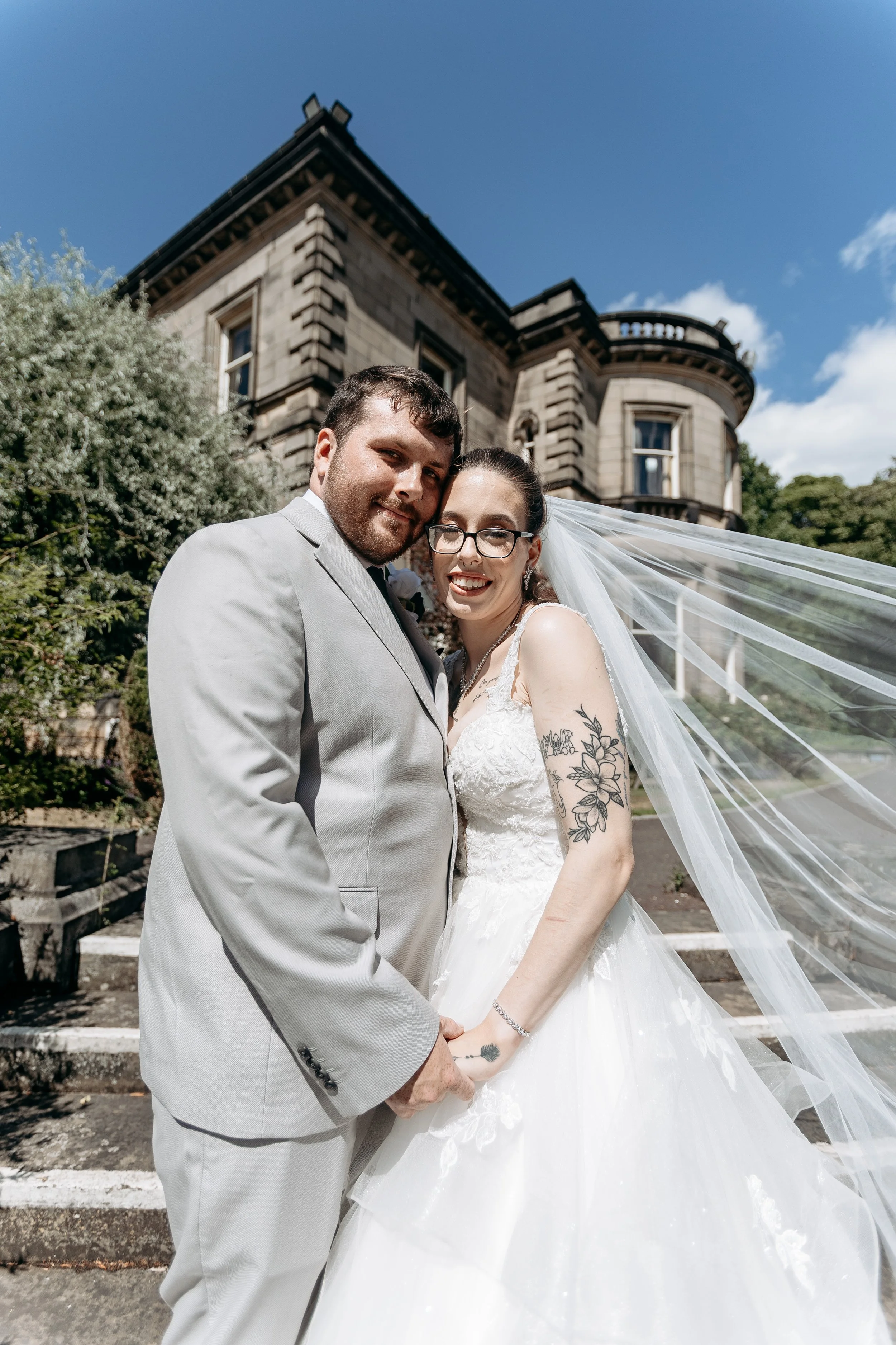 A newlywed couple in wedding attire standing outdoors in front of a large historic building during daytime. The groom is wearing a light gray suit, and the bride is in a white lace wedding dress with glasses and tattoos on her arms. They are holding 