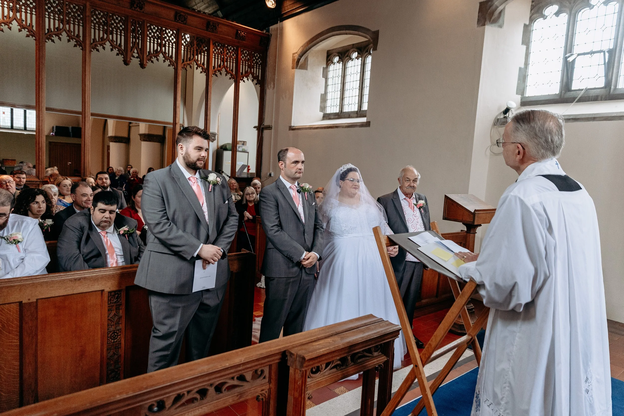 A bride and groom standing before a priest during their wedding ceremony inside a church, with guests seated in the background.