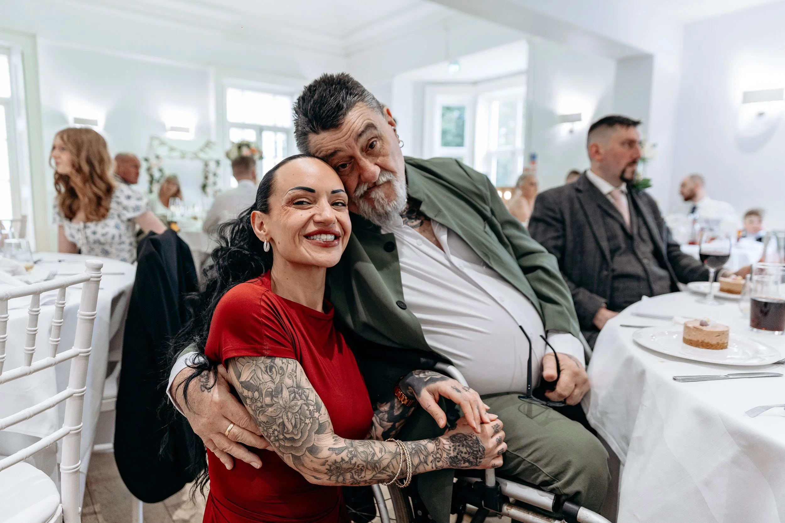 A happy woman with tattoos on her arms smiling and sitting close to a man in a wheelchair at a formal event, with others in the background at a banquet hall.