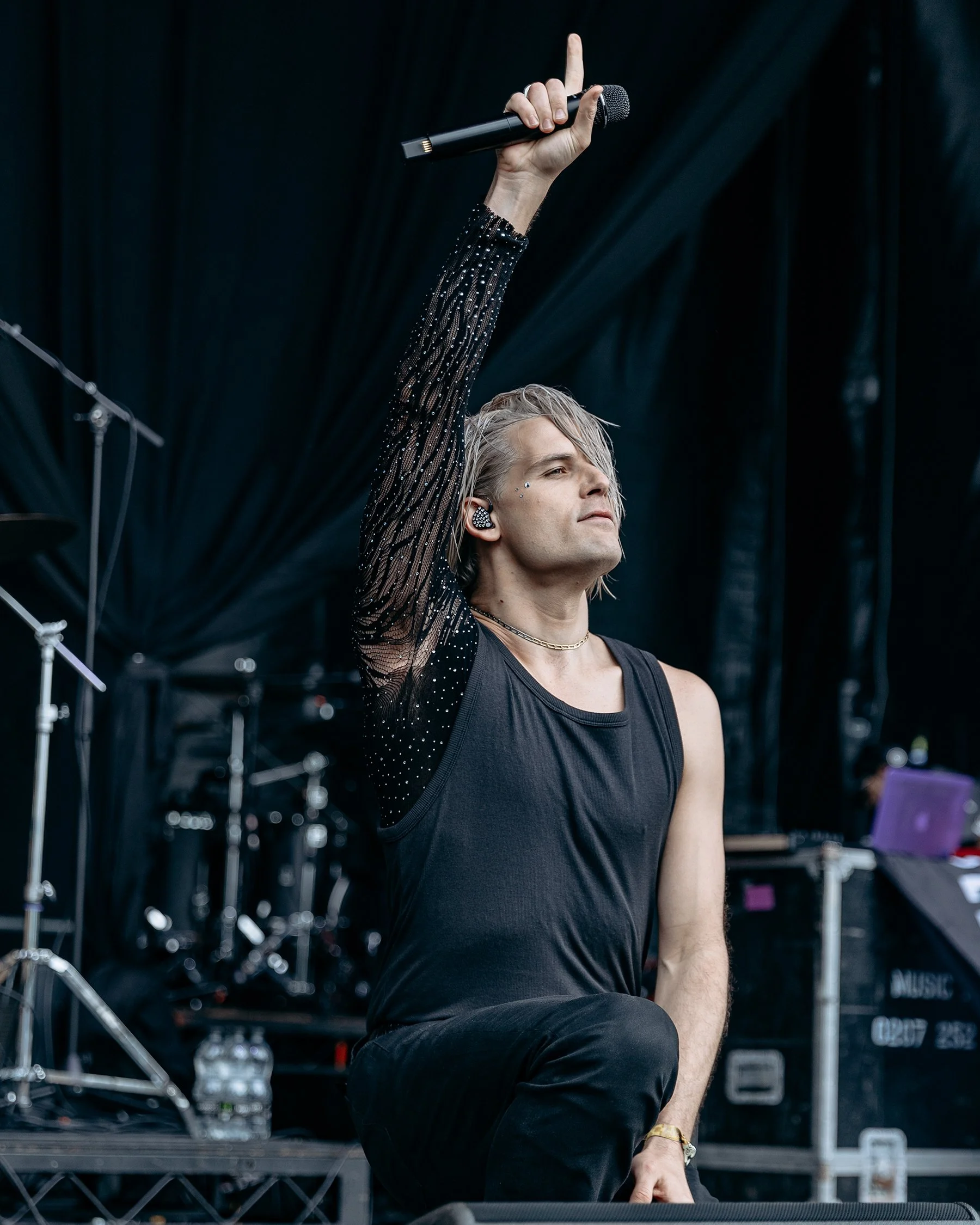 A male performer on stage holding a microphone in one hand with his arm raised, dressed in a black tank top and sheer black sleeves with rhinestones, performing at a music event.