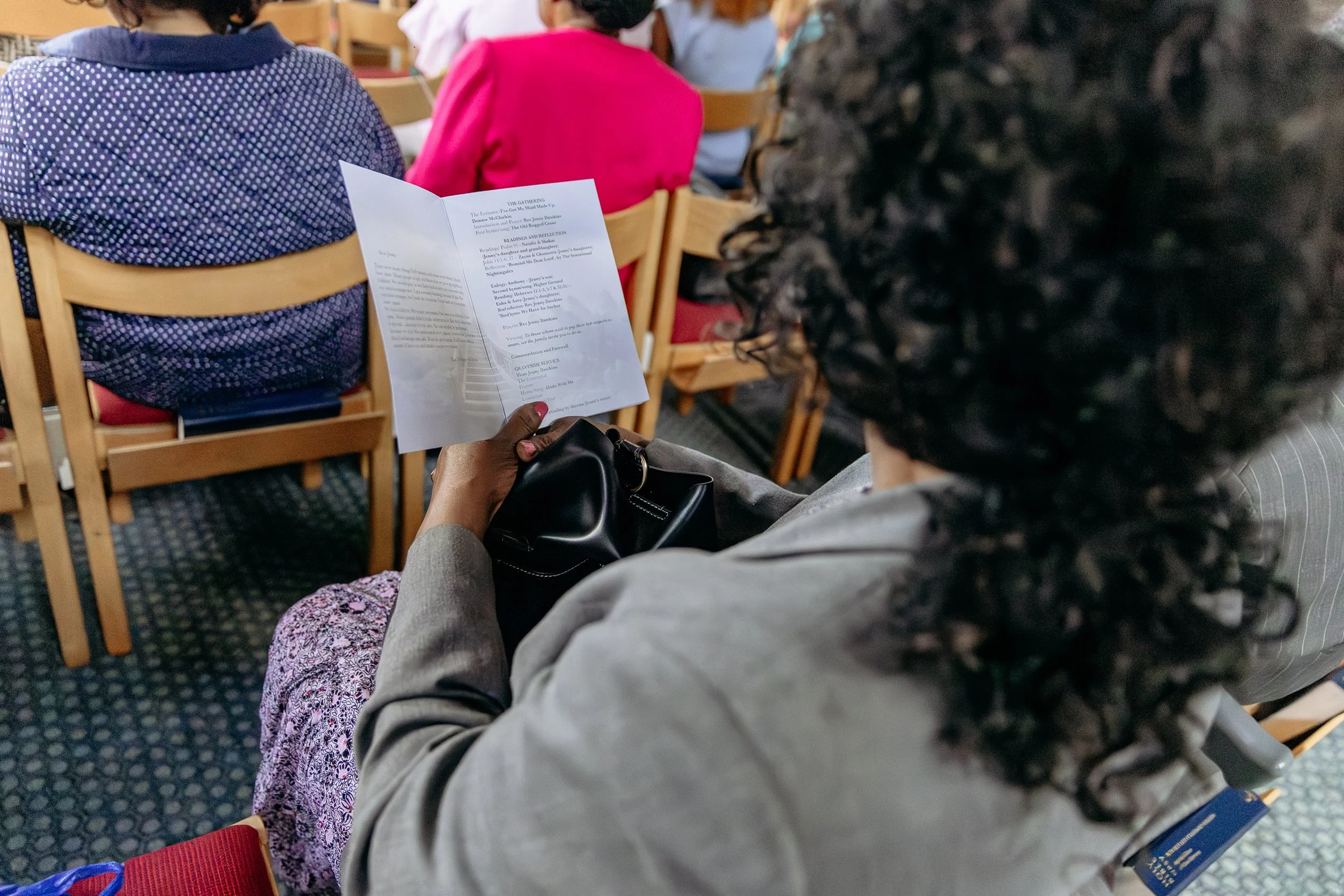 A woman with dark curly hair reading a program or booklet at a conference or seminar, seated among other attendees on wooden chairs.