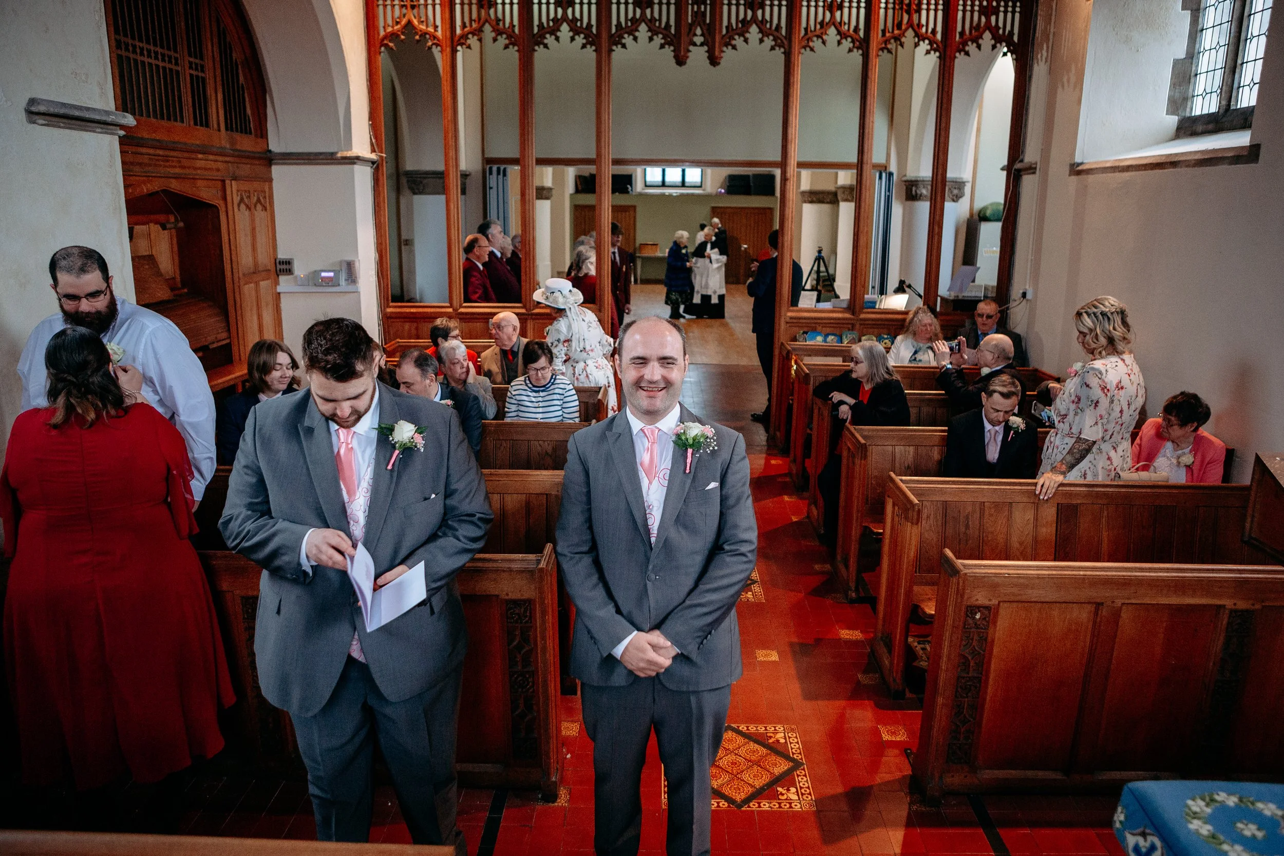 A wedding ceremony inside a church with guests seated in wooden pews. The groom stands at the front, smiling, wearing a gray suit with a pink tie and boutonniere. Other guests, dressed in formal attire, are talking and preparing for the ceremony.