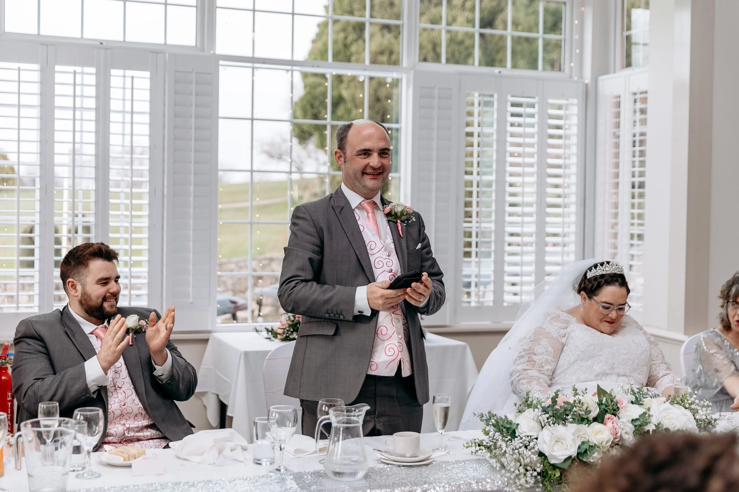 A wedding reception with a man in a suit making a speech, seated next to a bride in a wedding dress and tiara, and another man clapping nearby, inside a bright room with large windows.