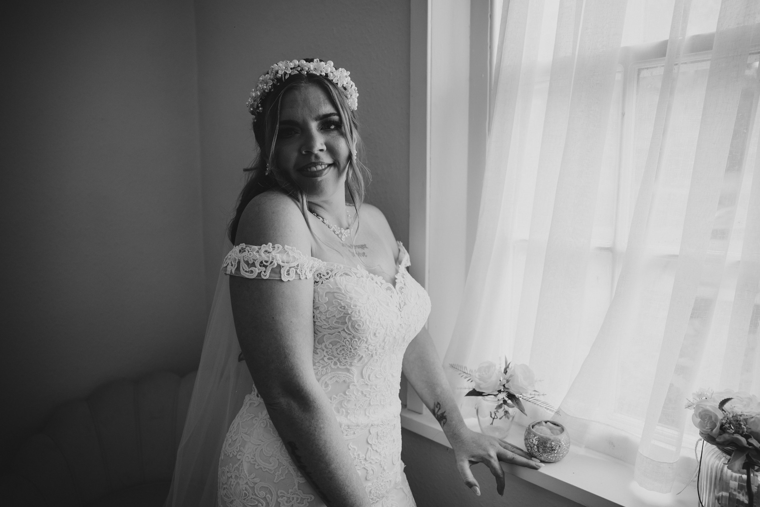 Black and white photo of a bride in a lace wedding dress, smiling, wearing a flower crown, standing by a window with sheer curtains.