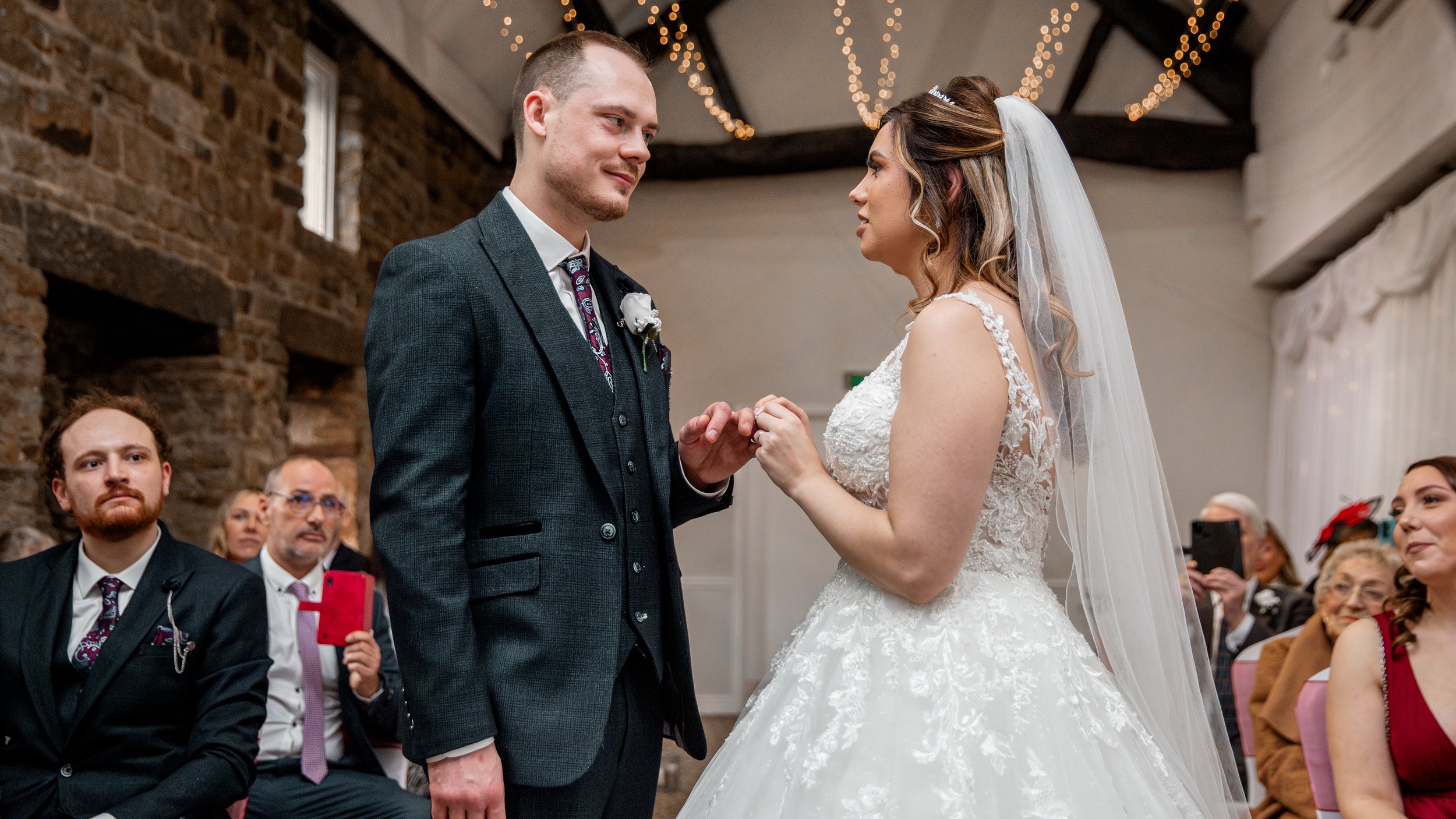 A bride and groom exchange rings during their wedding ceremony in a cozy indoor setting, with wedding guests watching.