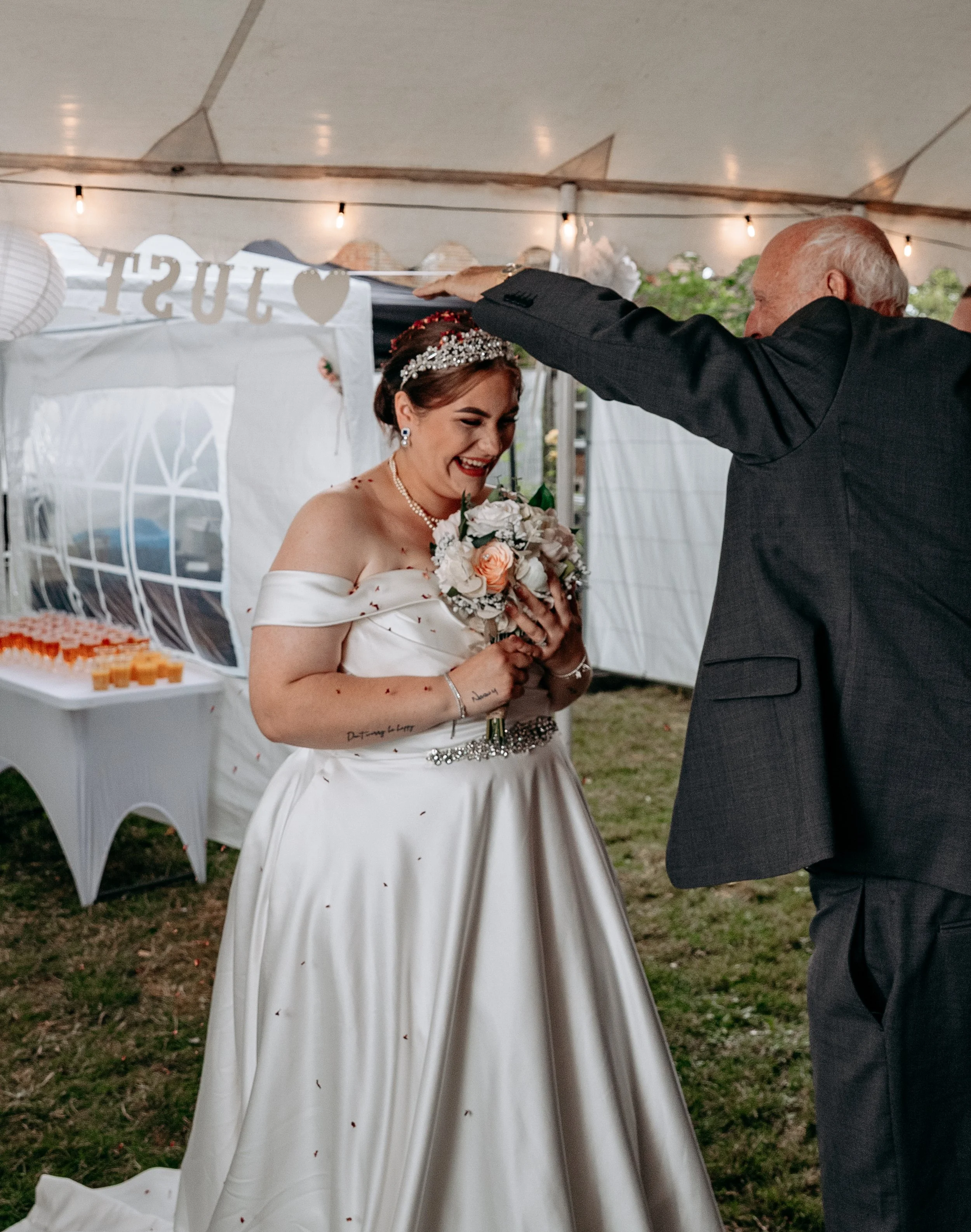 A bride in a white wedding dress holding a bouquet of flowers, smiling happily as a man touches her head in a celebration under a tent.