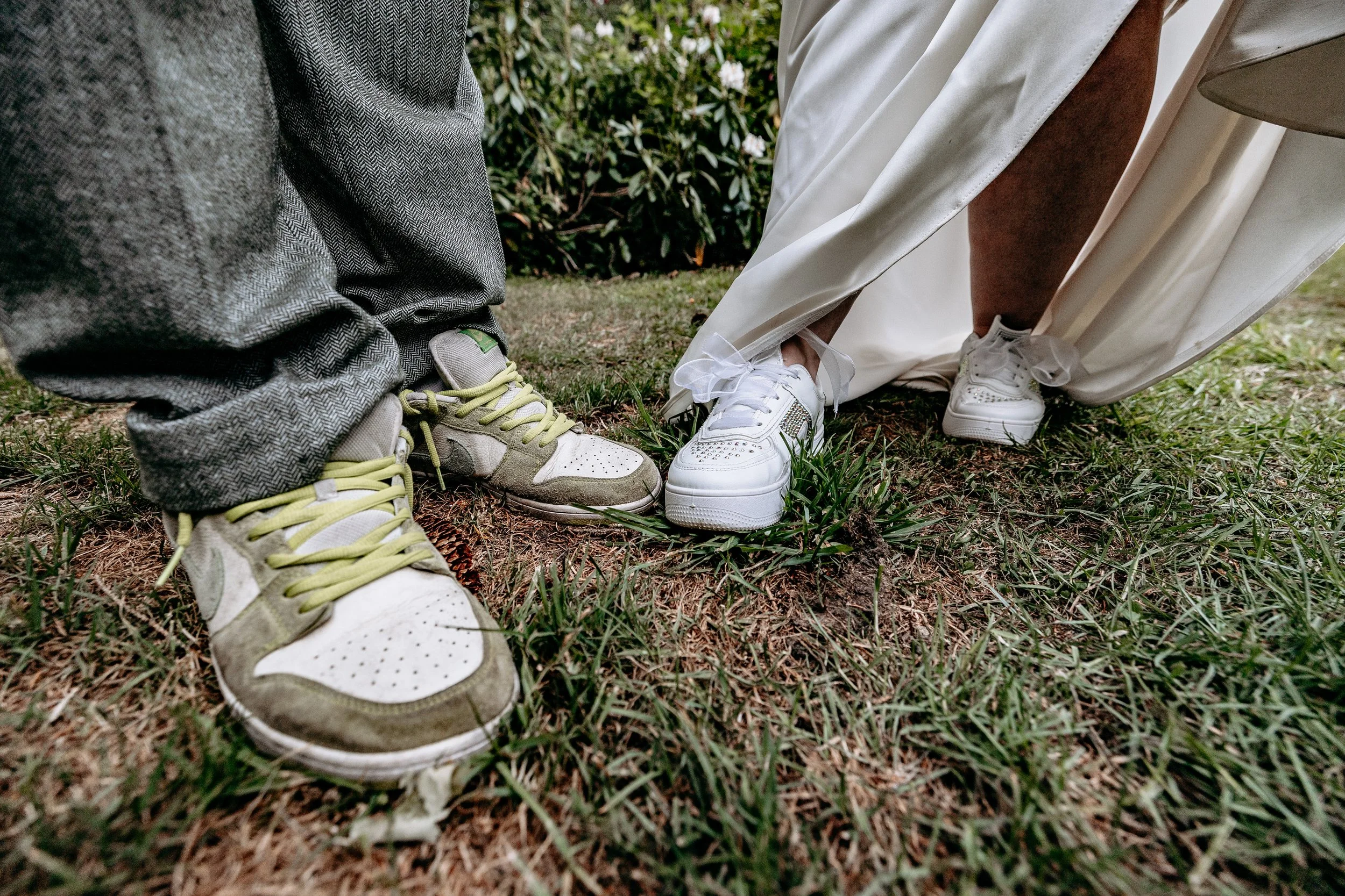 Close-up of the shoes of two people standing on grass, one wearing vintage sneakers and the other white sneakers with a long white dress and beige coat.