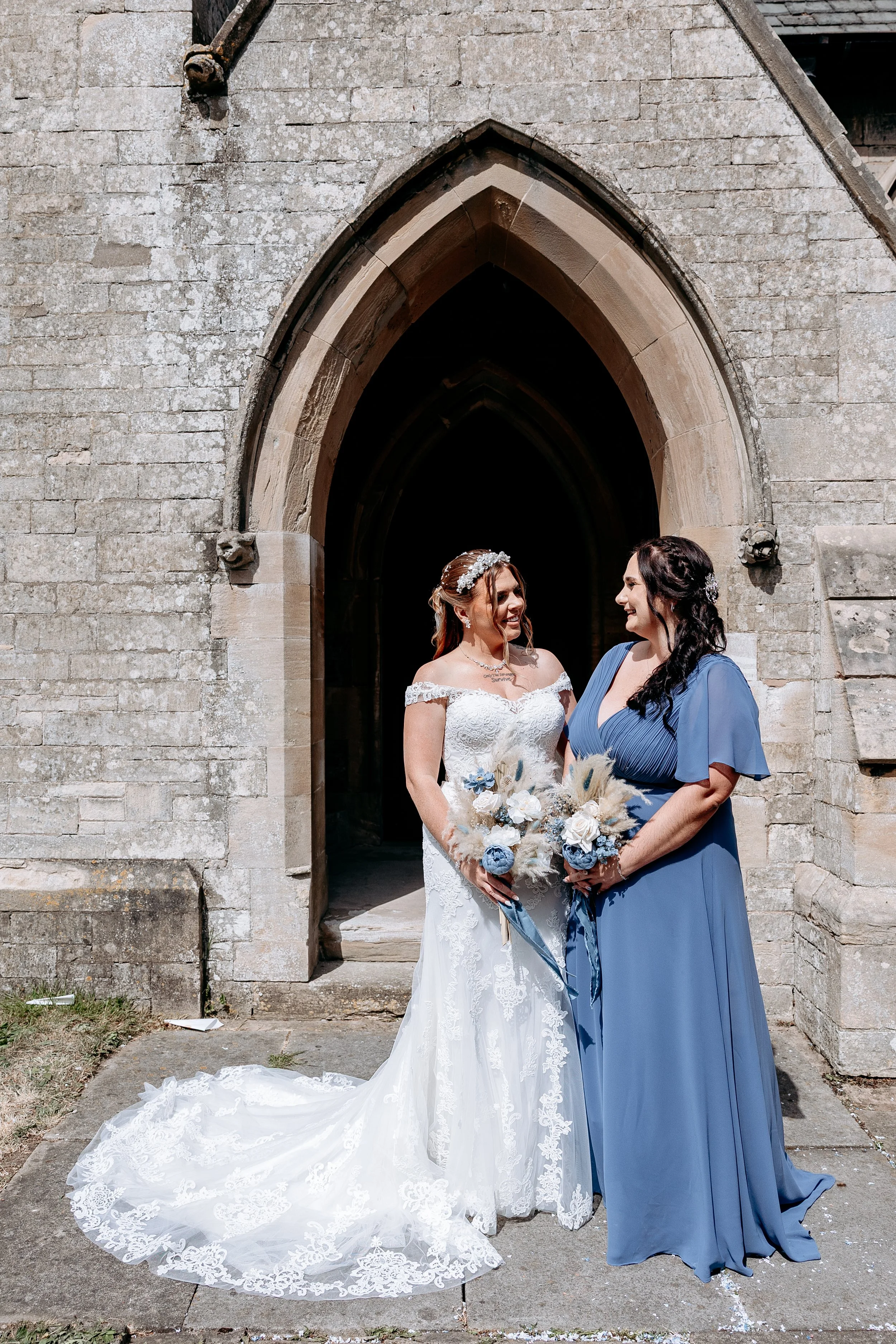 Two women, a bride in a white lace wedding dress and a bridesmaid in a blue dress, standing outside a stone building with an arched doorway, holding bouquets, smiling at each other.
