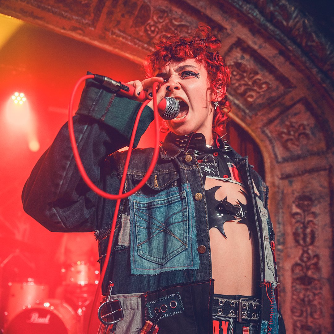 A woman with curly red hair singing into a microphone on stage, wearing a black denim jacket with colorful patches, a black bat-shaped chest accessory, and bold makeup.