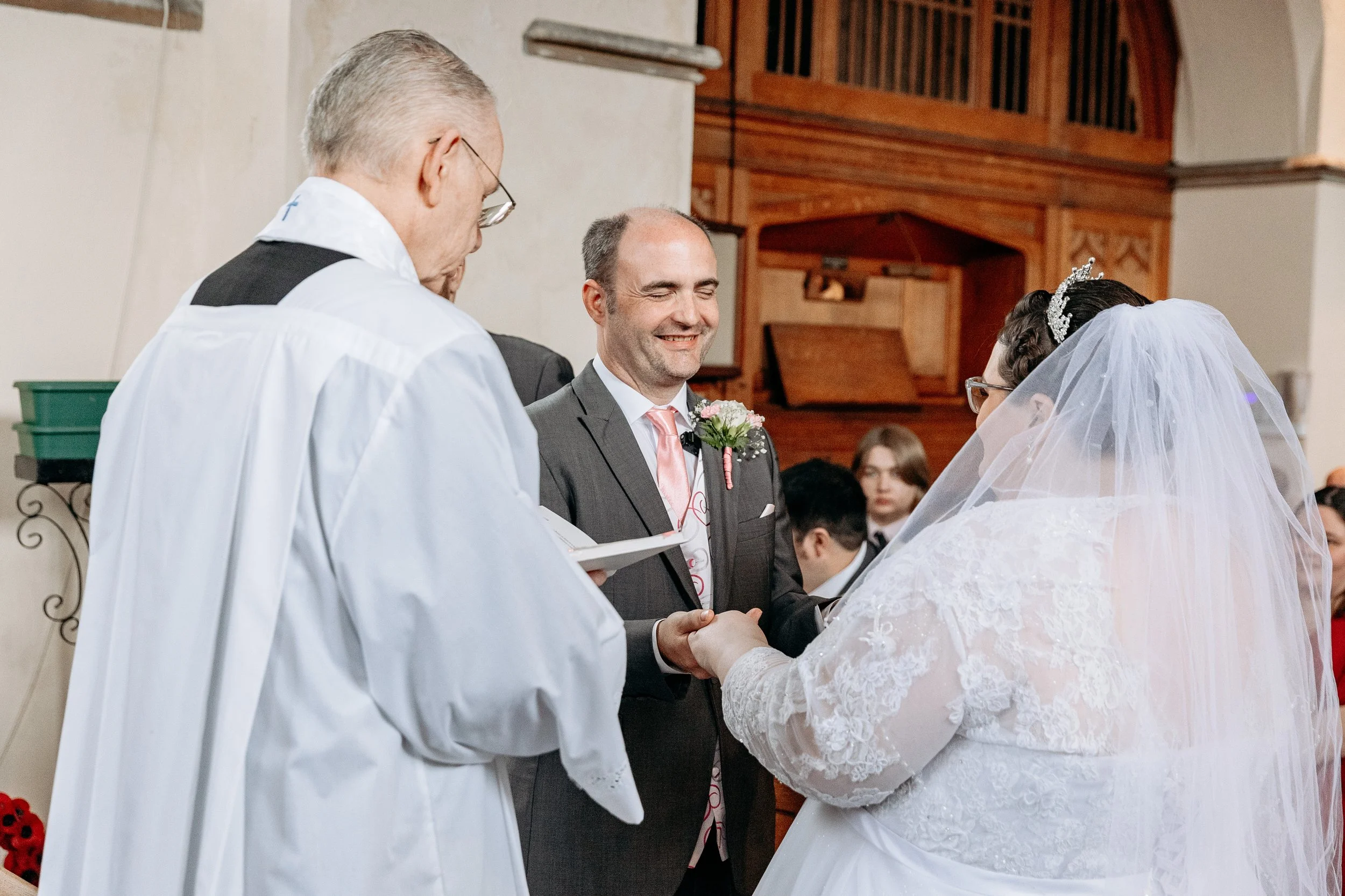 A wedding ceremony with a bride and groom holding hands, an officiant reading from a book, and guests watching in a church.
