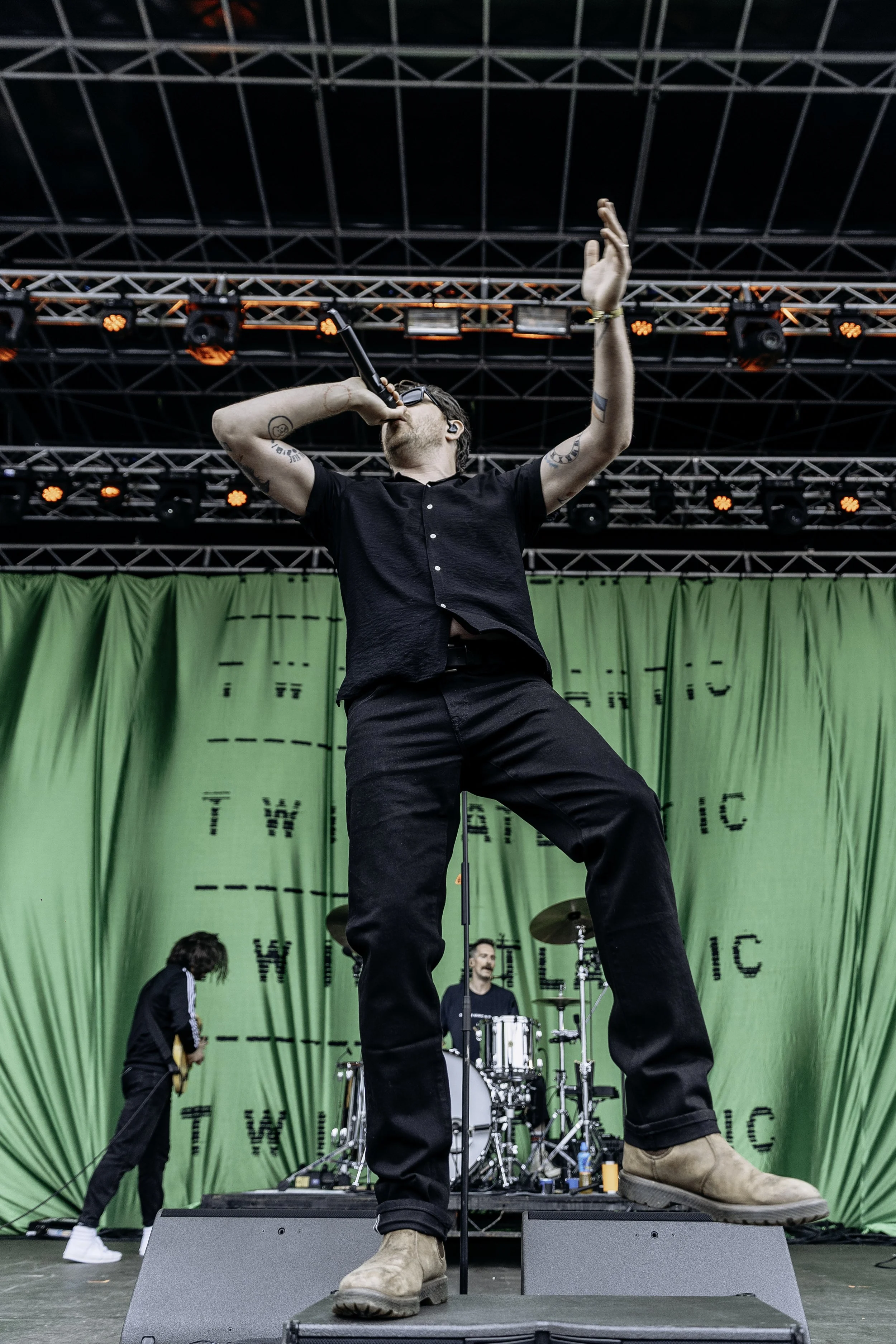A male singer with tattoos, wearing a black shirt and beige boots, is performing on stage with a microphone, raising one hand. A drummer and a guitarist are visible in the background, with a green curtain and stage lighting above.
