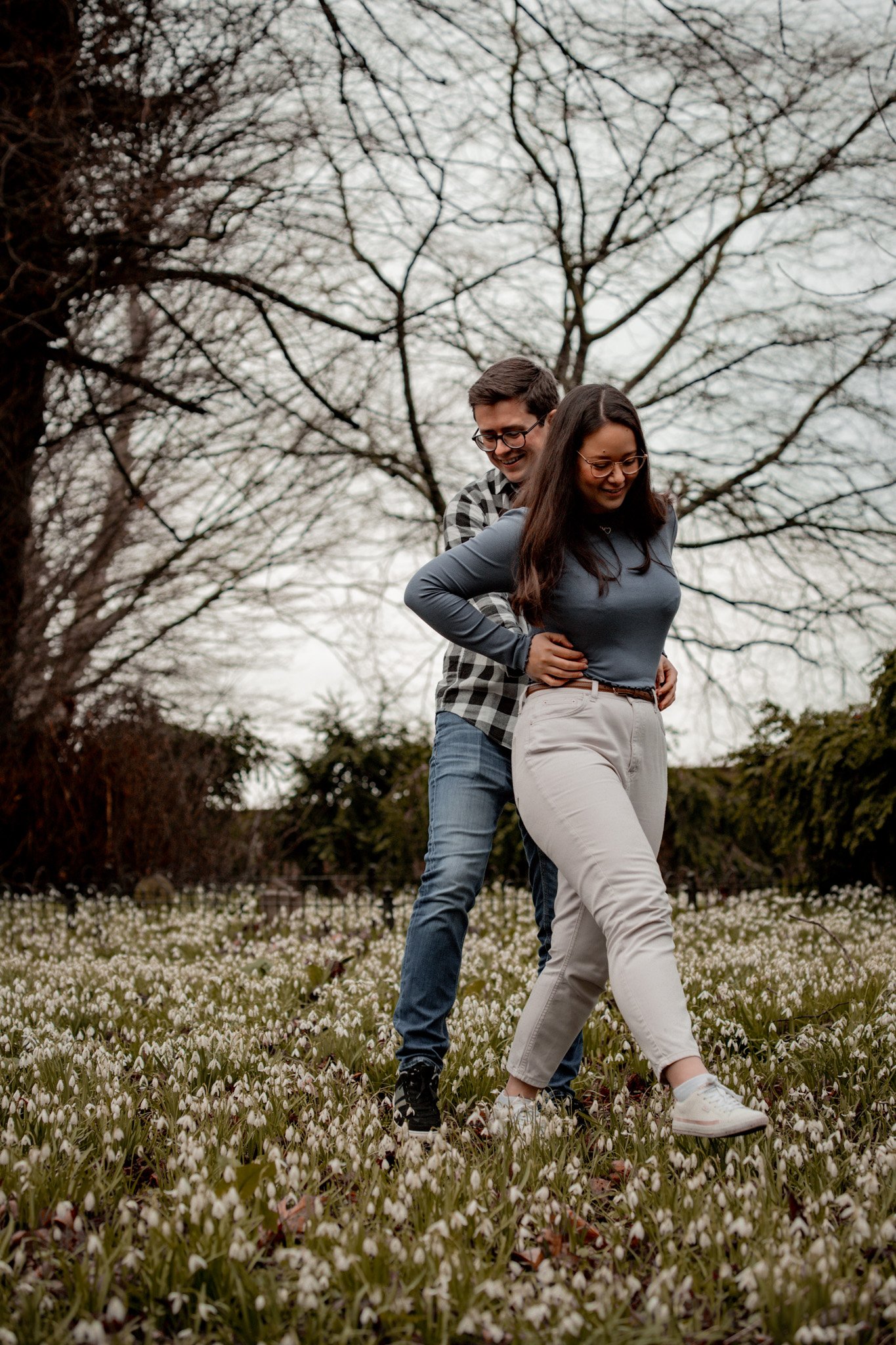 A man and woman walking together in a field of small white flowers, with trees and an overcast sky in the background.