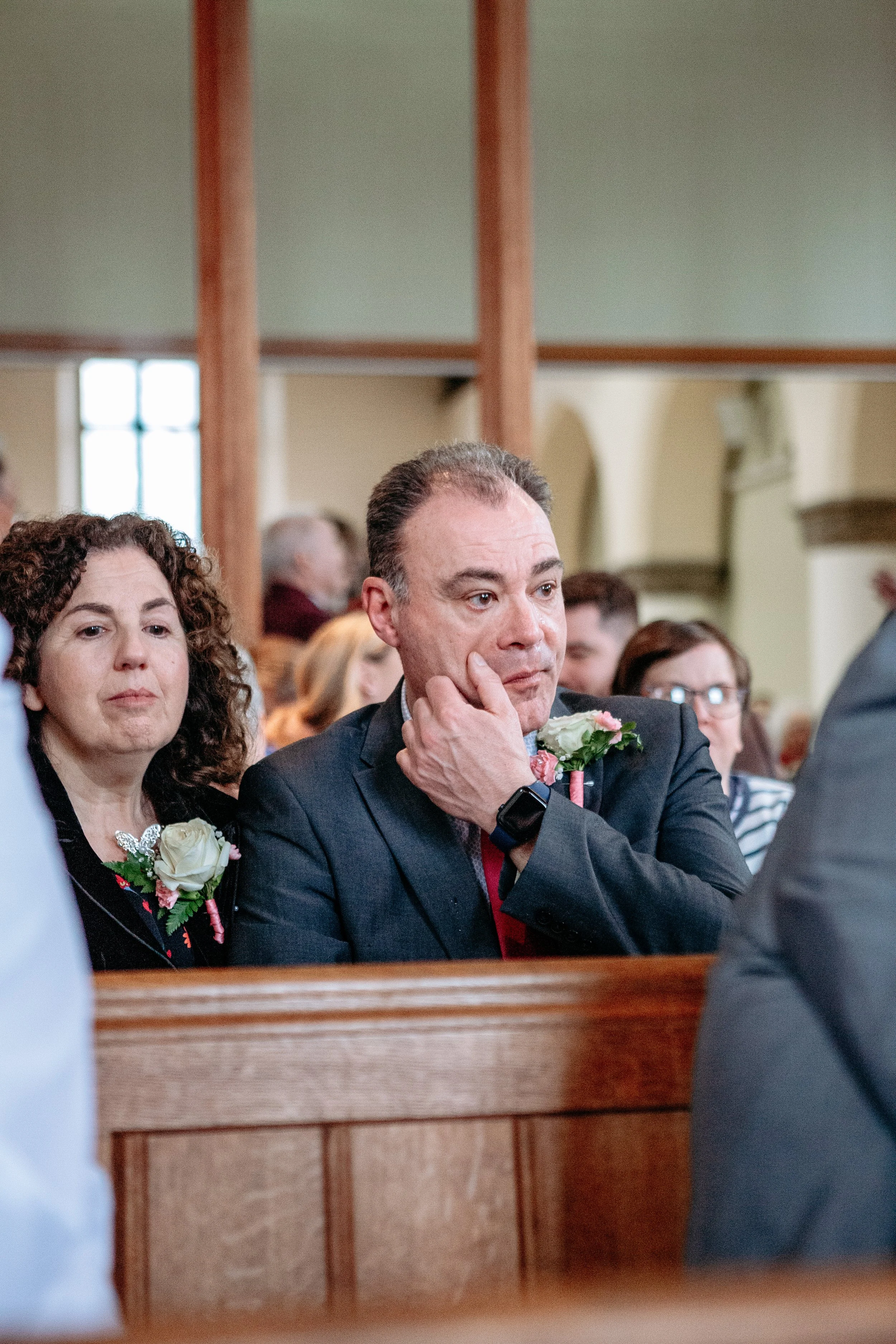 A man in a suit sitting in a church pew during a wedding ceremony, appearing emotional with his hand on his chin, surrounded by other guests.