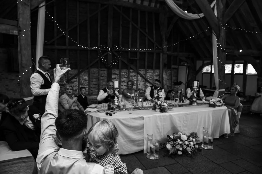 A black and white photo of a wedding reception in a barn with a long table. Guests are seated and a man is standing, holding a glass. Decorations include string lights and floral arrangements.