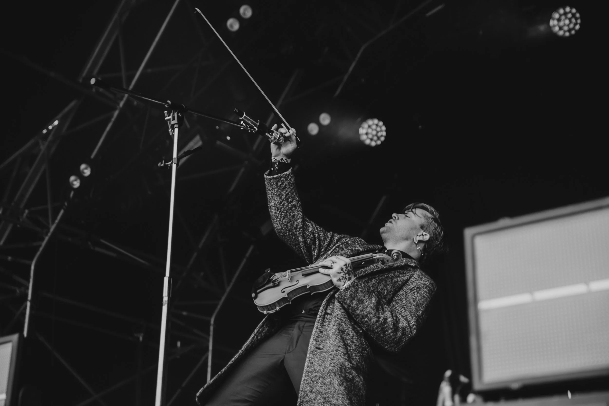 A musician on stage holding a violin and raising a bow, wearing a coat with stage lights and equipment in the background.
