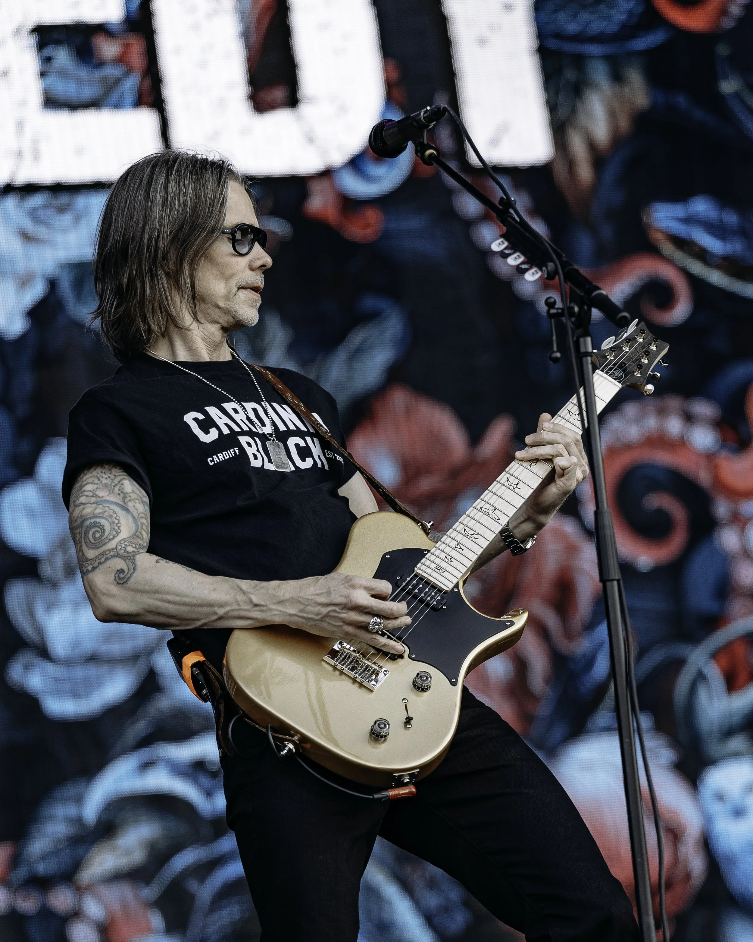 A male musician with shoulder-length hair, wearing sunglasses and a black t-shirt, playing an electric guitar on stage with a large, colorful abstract background.