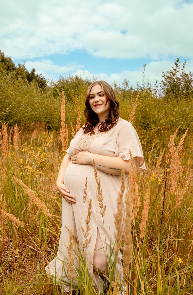 Pregnant woman standing in a grassy field with wildflowers, smiling and holding her belly, under a partly cloudy sky.