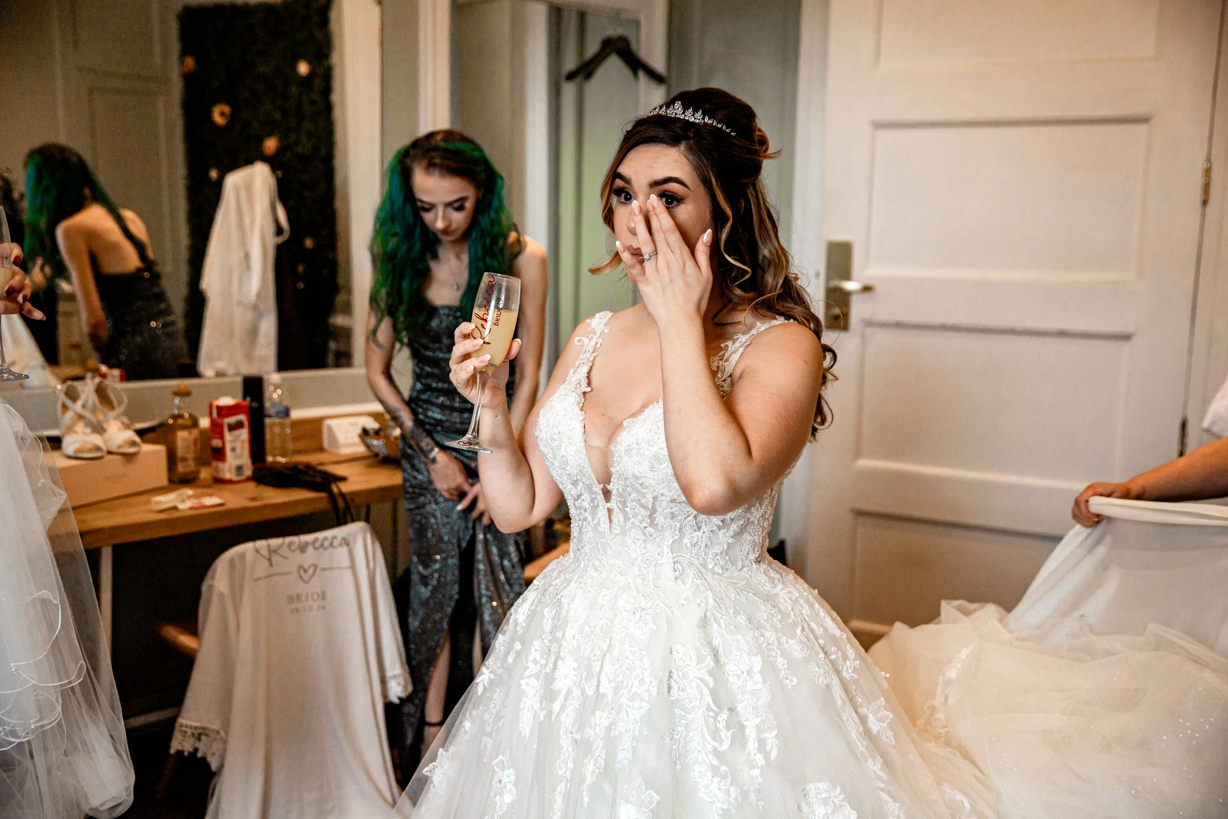 A bride in a wedding dress holding a champagne glass with tears of joy, while her friends prepare in a dressing room.