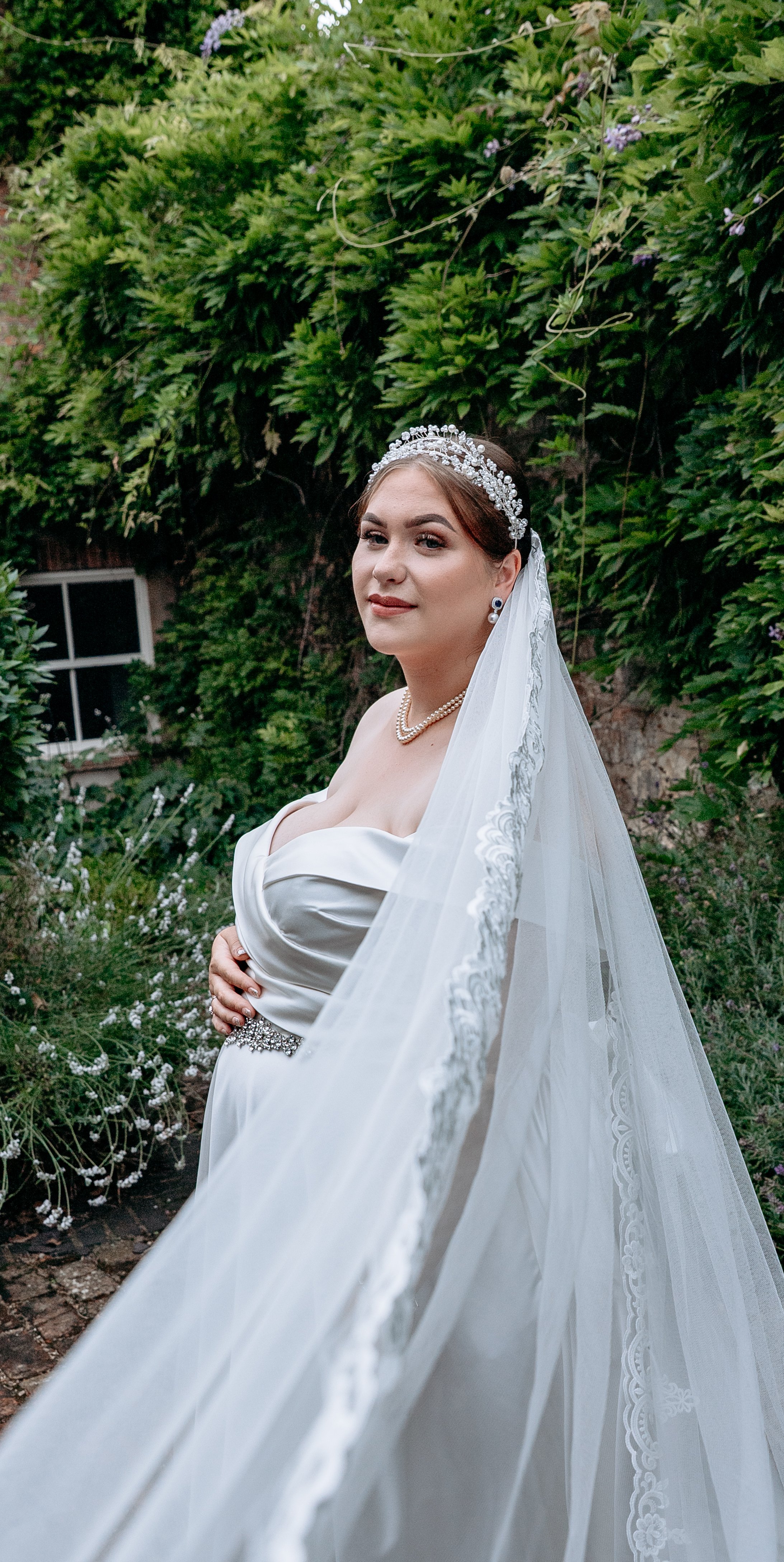 Bride in a white wedding dress with a lace veil and pearl jewelry standing outdoors with greenery in the background.