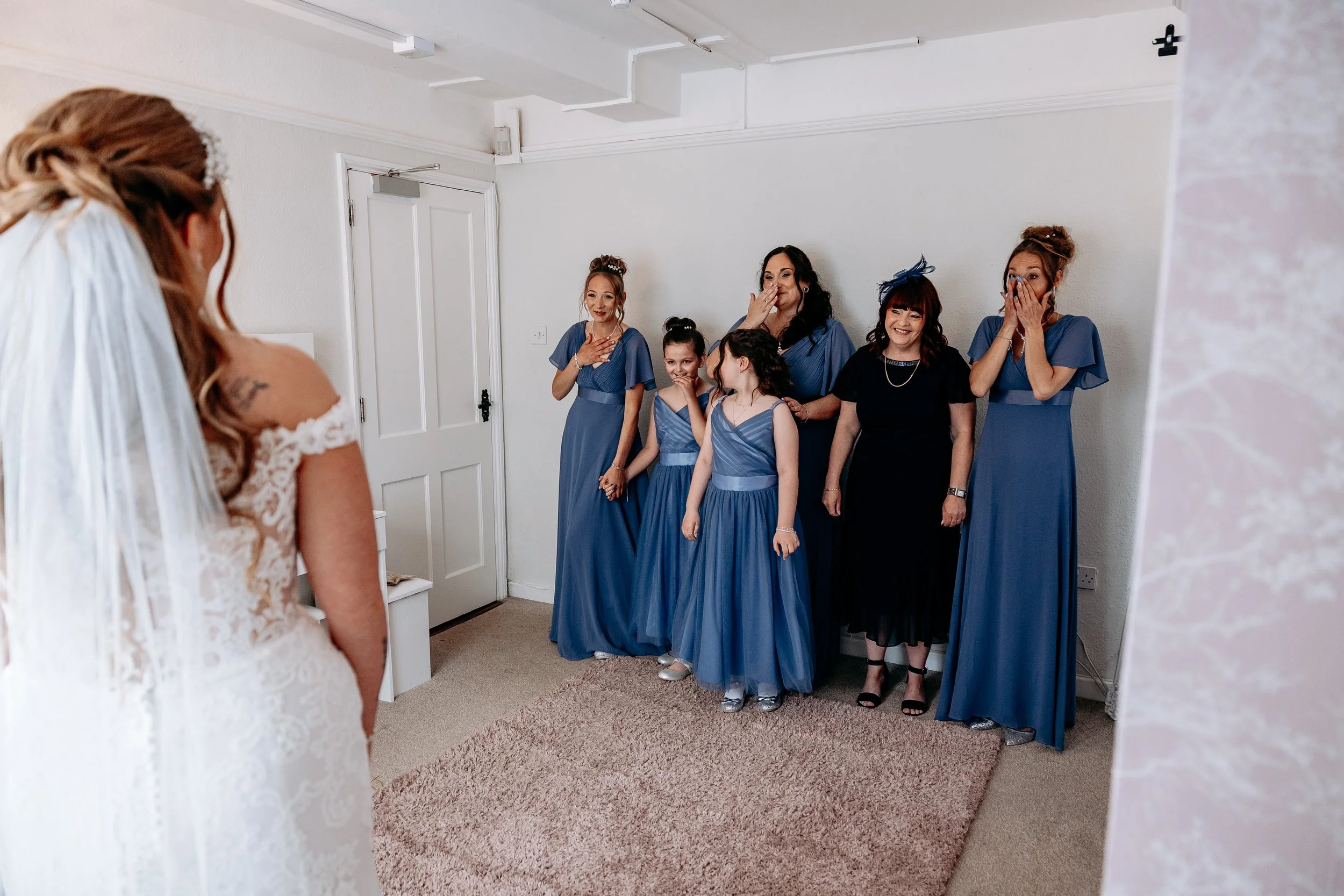 A bride in a white lace wedding dress looking at six women, including young girls and adults, dressed in matching blue dresses, with some showing emotional reactions, in a room before a wedding ceremony.