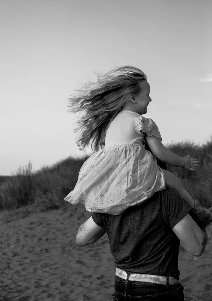 Black and white image of a child sitting on an adult's shoulders, walking on a sandy path with grass in the background. The child is wearing a dress and has long hair blowing in the wind.