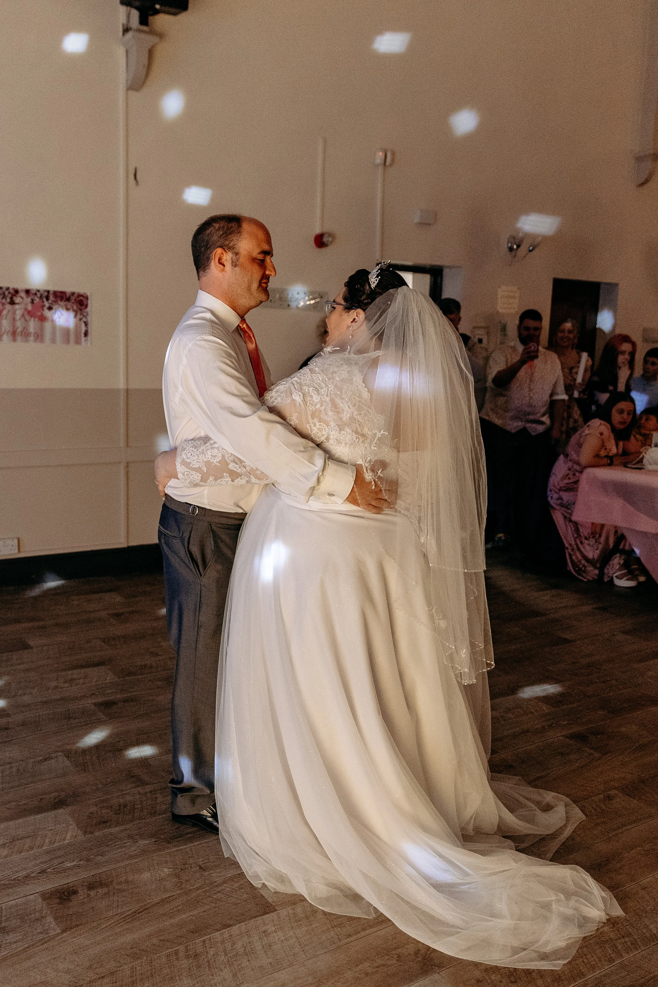 A bride and groom dancing at their wedding reception, surrounded by guests, with a dimly lit atmosphere.