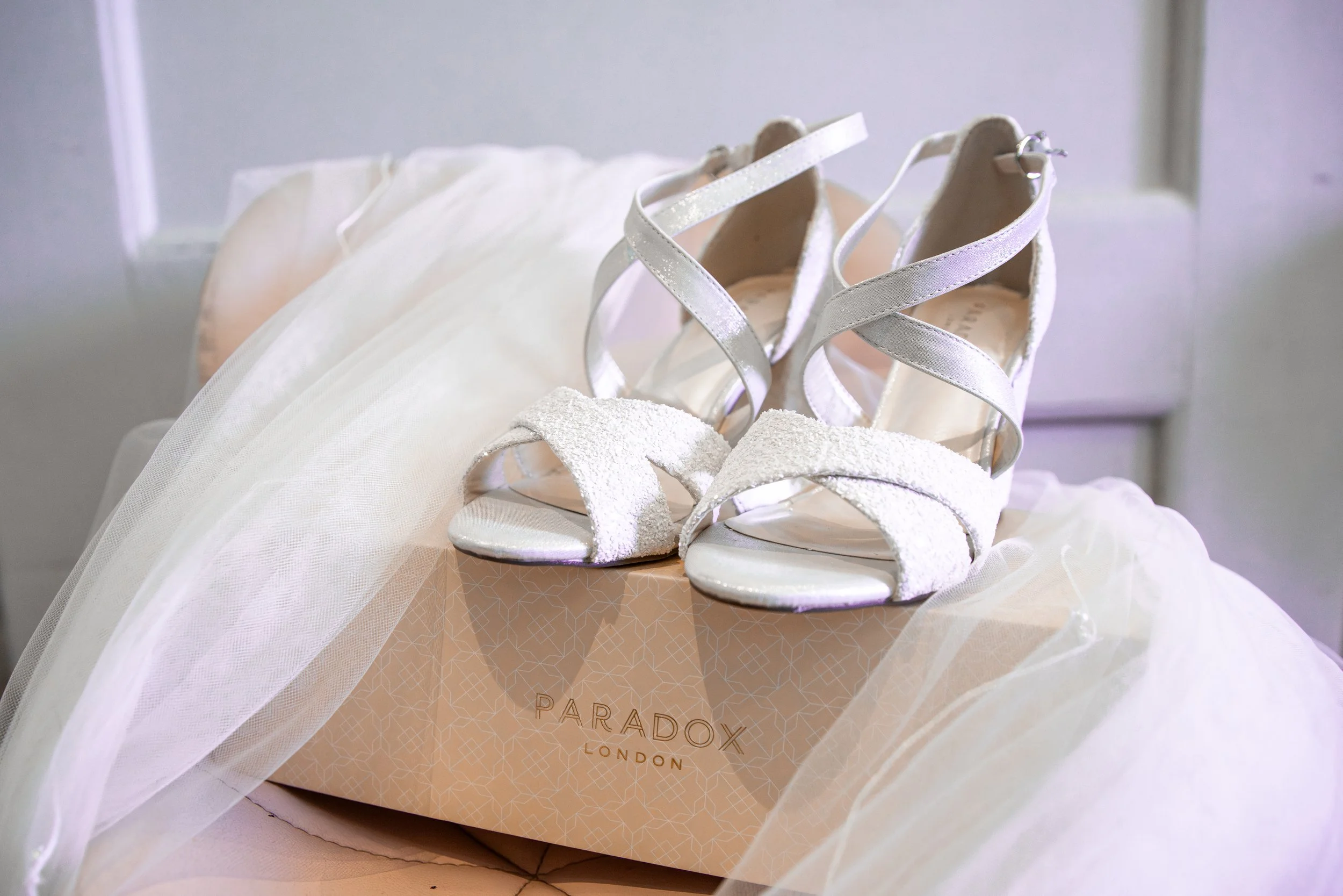 A pair of white, glittery high-heeled sandals resting on a pink box labeled 'Paradox London,' draped with a sheer, white fabric that resembles a wedding veil.