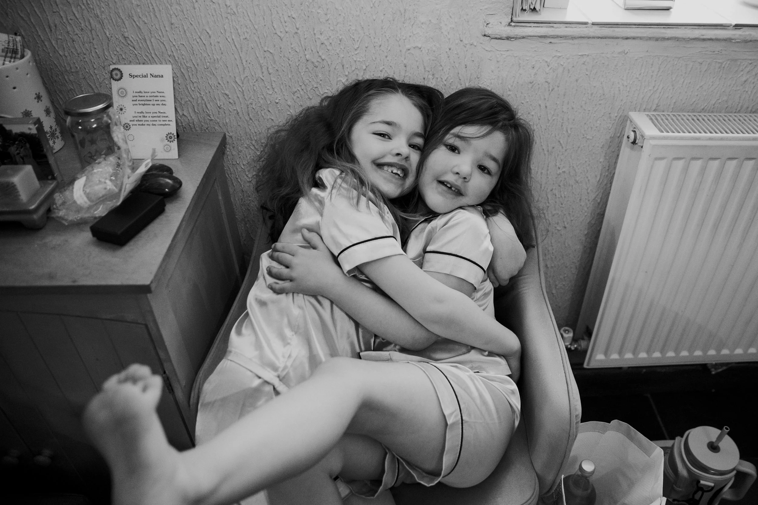 Two young girls hugging each other on a chair, smiling, in a cozy room with a textured wall and a radiator, in black and white.