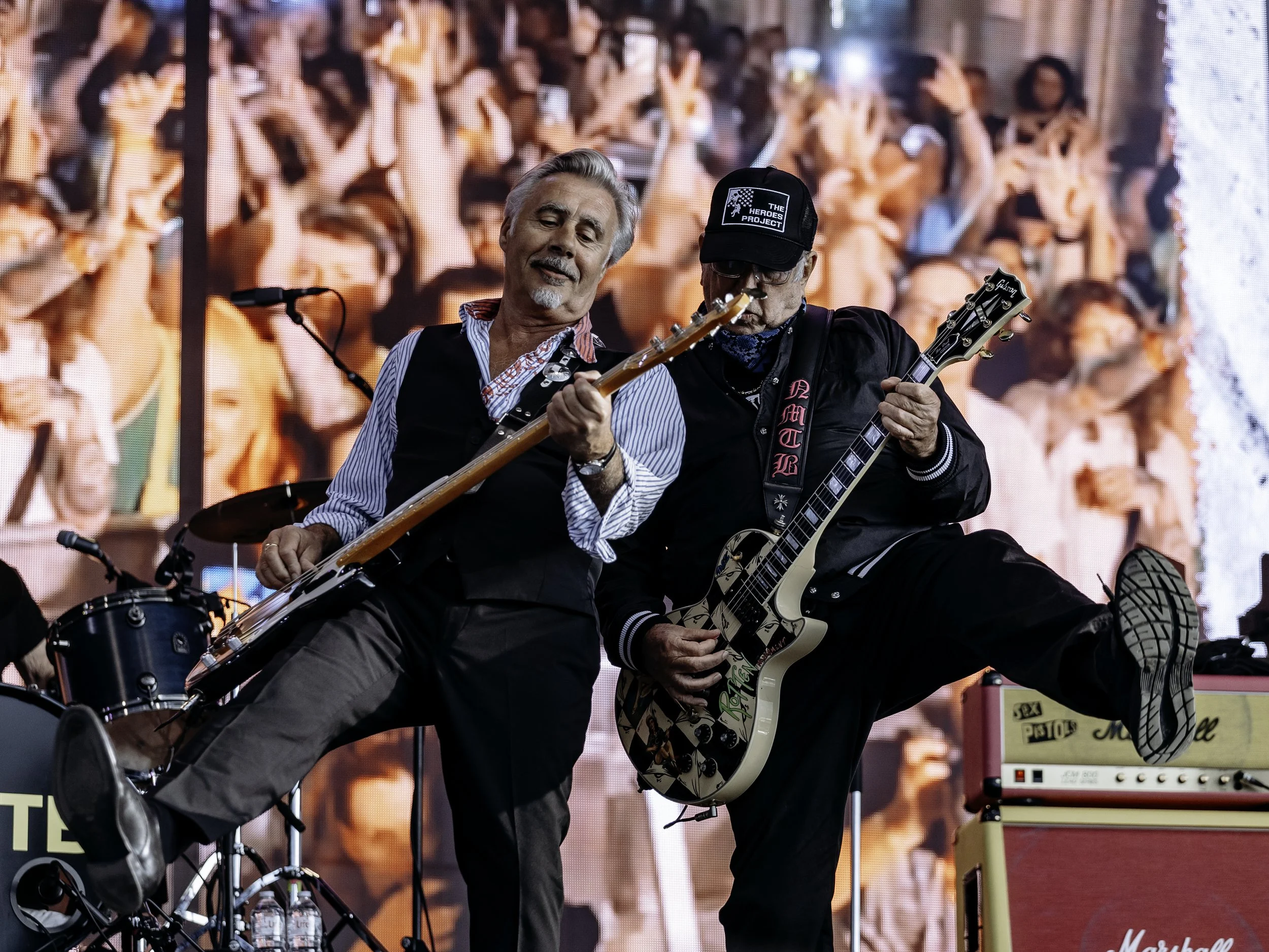 Two male musicians playing electric guitars on stage during a live concert, with a large crowd visible on a screen behind them.
